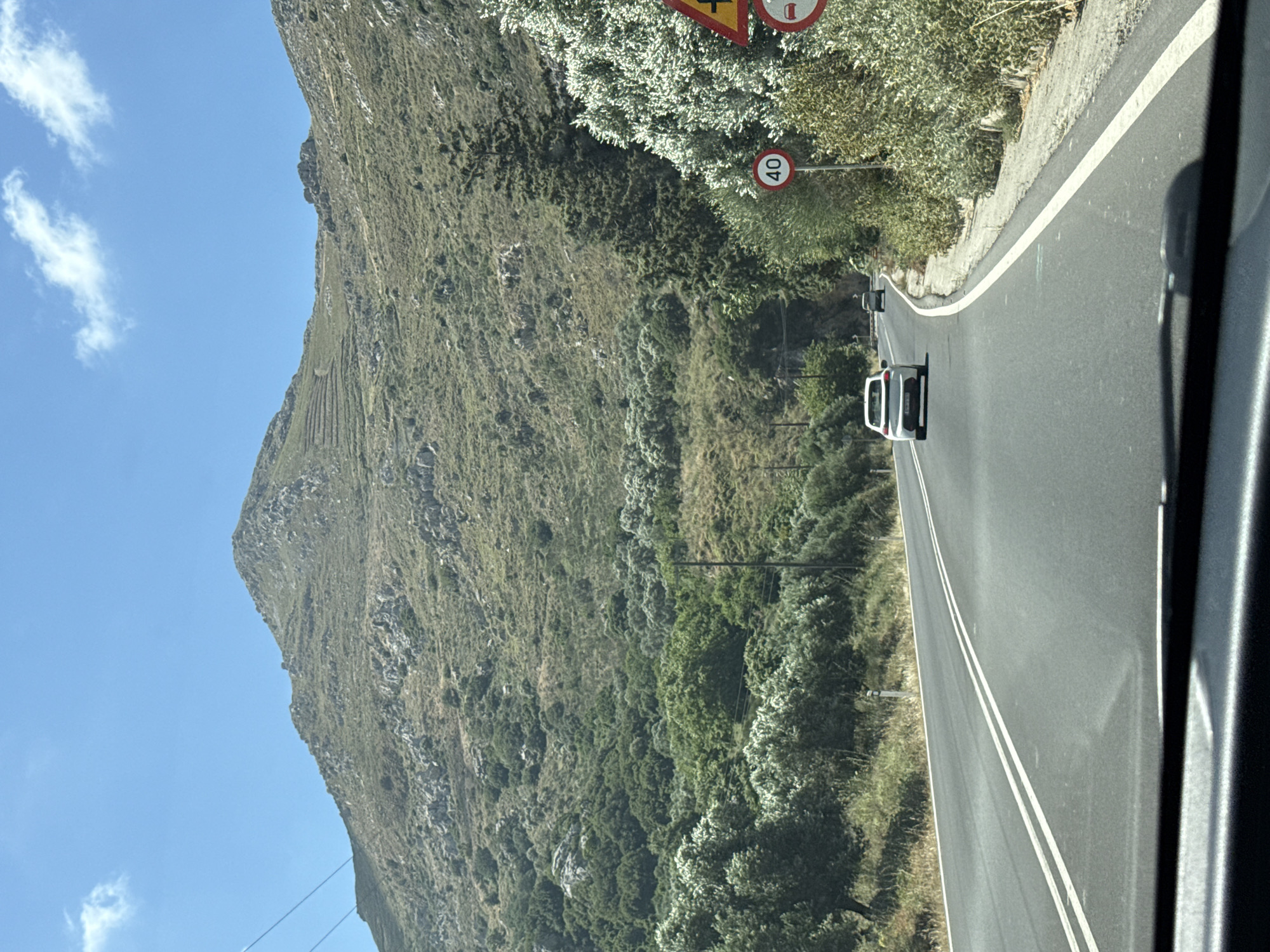 An eye-level outdoor shot shows a curved asphalt road with a white double line in the center, leading into a mountainous landscape under a bright blue sky with scattered white clouds. On the right side of the road, there is a roadside with dense green foliage and a white-lined embankment. Traffic signs are visible on the right, including a circular sign with "40" in red and a yellow triangular sign with a red border. Two cars are seen on the road; a white car is in the foreground, and a darker car is further ahead. The road is bordered by vegetation on both sides, with trees and shrubs creating a naturalistic setting. The background is dominated by a large, barren mountain with visible terraces and patches of dry grass and rocks. Electrical poles and wires are present along the left side of the road, hinting at the presence of infrastructure. The shot appears to be taken from a moving vehicle, as indicated by the blurred foreground elements and the perspective of the road.