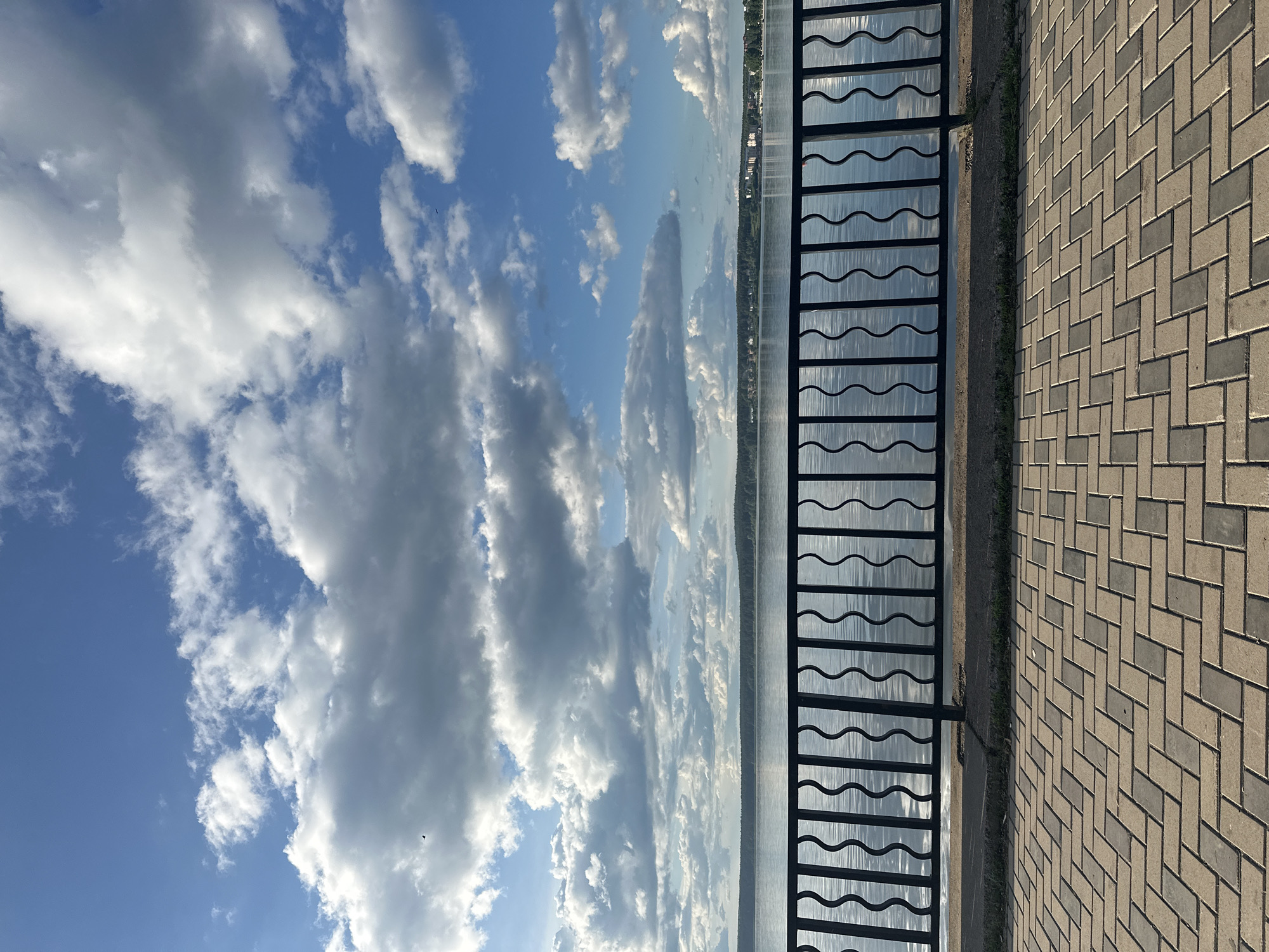 The image depicts a section of the Votkinsk embankment in Russia, showing a paved pedestrian walkway to the right, separated from the water by a black metal railing with a wavy design. Beyond the railing is a body of water reflecting the sky, with a distant shoreline visible. The sky is predominantly blue, filled with numerous puffy white clouds. The image is oriented sideways.