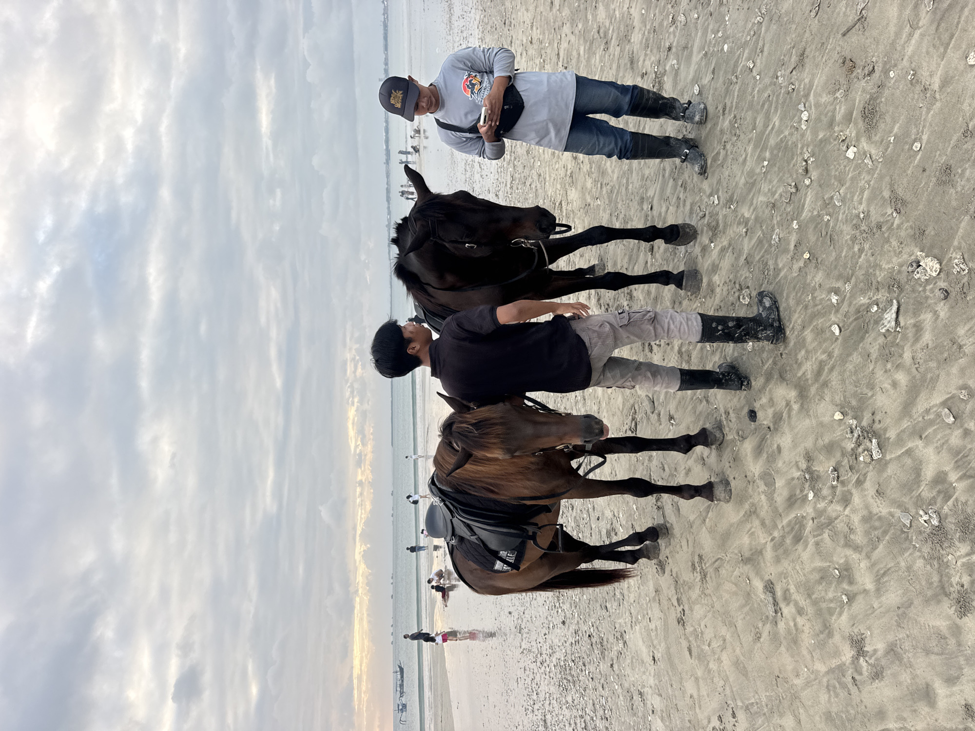 This is a photograph of two horses and two young men on a sandy beach in Kuta Selatan, Indonesia. The sky is overcast with some patchy clouds. The ocean is in the background, with several boats visible on the water. There are a few other people in the distance on the beach and in the water. One horse, a chestnut color, is equipped with a saddle. The other horse is a dark brown or black color and is bridled. Both horses are standing on the wet sand near the water's edge. The two men are standing close to the horses. The man on the left is wearing a black t-shirt and grey cargo pants. The man on the right is wearing a grey long-sleeved shirt, blue jeans, and black boots. He is holding a mobile phone in his hand and looking at it. The scene appears to be set during a cloudy day, possibly early morning or late afternoon.