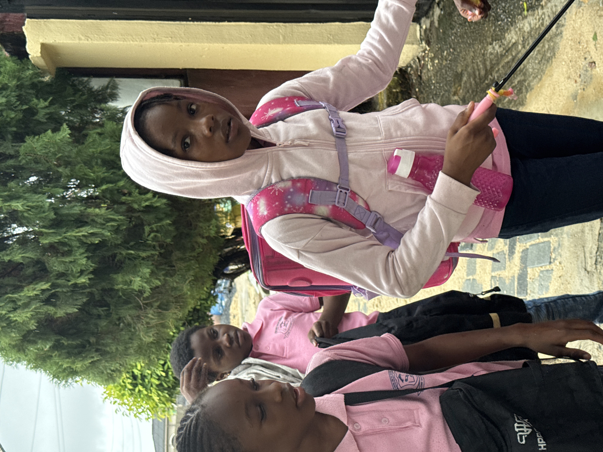 A group of children are standing outside a building. The child in the foreground is wearing a pink hooded sweatshirt, a backpack, and holding a pink water bottle and a small fishing rod. Two other children wearing pink shirts are in the background. The location is Ado, Nigeria.