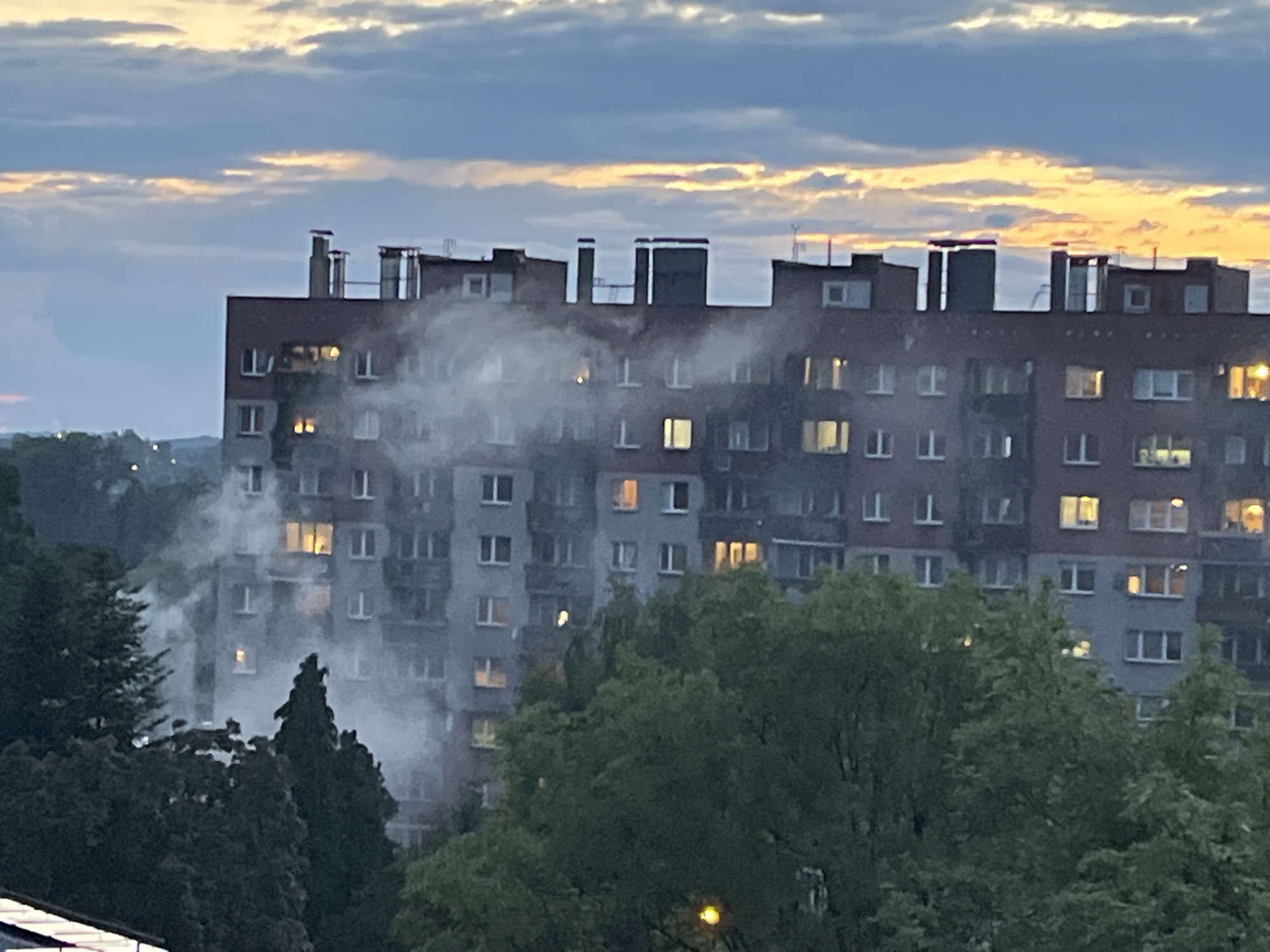 An outdoor medium shot captures a residential building at dusk, possibly in a city. The building, which appears to be an apartment complex with multiple stories and windows, is partially obscured by billowing white smoke. Some windows on the building are illuminated with warm yellow light, suggesting that some residents are home or that lights are on. In the foreground, a dense canopy of green trees partially blocks the view of the lower portion of the building. The sky above is a gradient of dusky blues and grays, with streaks of soft orange and yellow light indicating the setting sun or twilight. The overall impression is one of an unusual event occurring in a residential area during the evening hours.