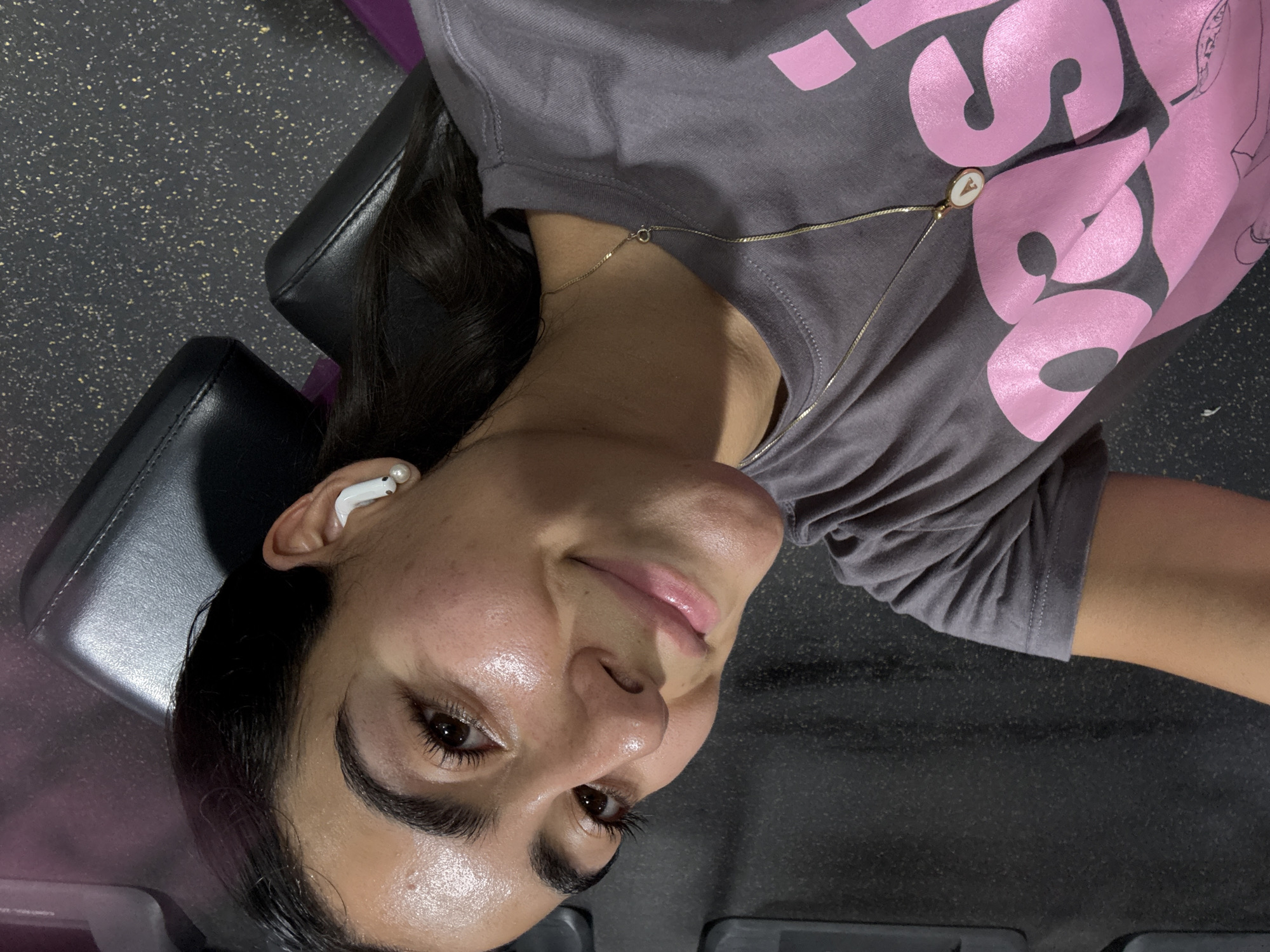 A woman is lying on a black padded bench in what appears to be a gym. She is fair-skinned with dark hair and is wearing a grey t-shirt with pink lettering and a gold necklace. She has an earbud in her left ear. The floor has a dark, speckled pattern. No further information regarding the location or city of Puebla, Mexico is evident in the image.