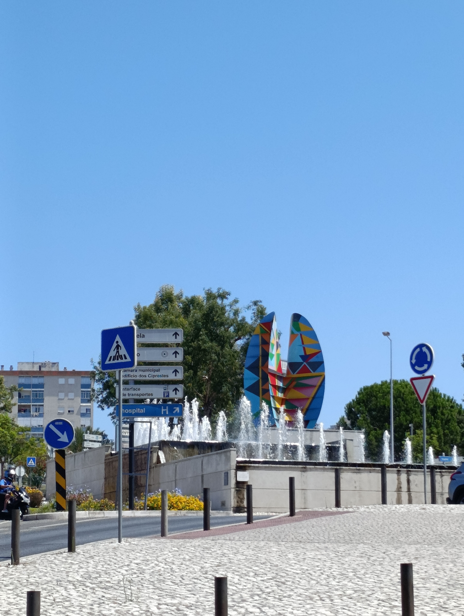 The image captures a street scene in Setúbal, Portugal, dominated by a vibrant, abstract sculpture located in the center of a roundabout with a fountain. The sculpture, composed of multi-colored, geometric shapes, is surrounded by multiple water jets. To the left of the sculpture, a pedestrian crossing sign is visible, along with directional signs indicating "Câmara municipal," "Edifício dos Ciprestes," "Interface de transportes," and "Hospital," with accompanying arrows and a parking symbol. A motorcyclist, wearing a helmet, is seen on the left side of the image, approaching the roundabout. In the background, a multi-story residential building with balconies and windows stands under a clear blue sky. A smaller, triangular red and white sign, indicating a yield or warning, is positioned near the roundabout signpost. The ground in the foreground is paved with cobblestones. The overall environment suggests an urban setting on a sunny day.