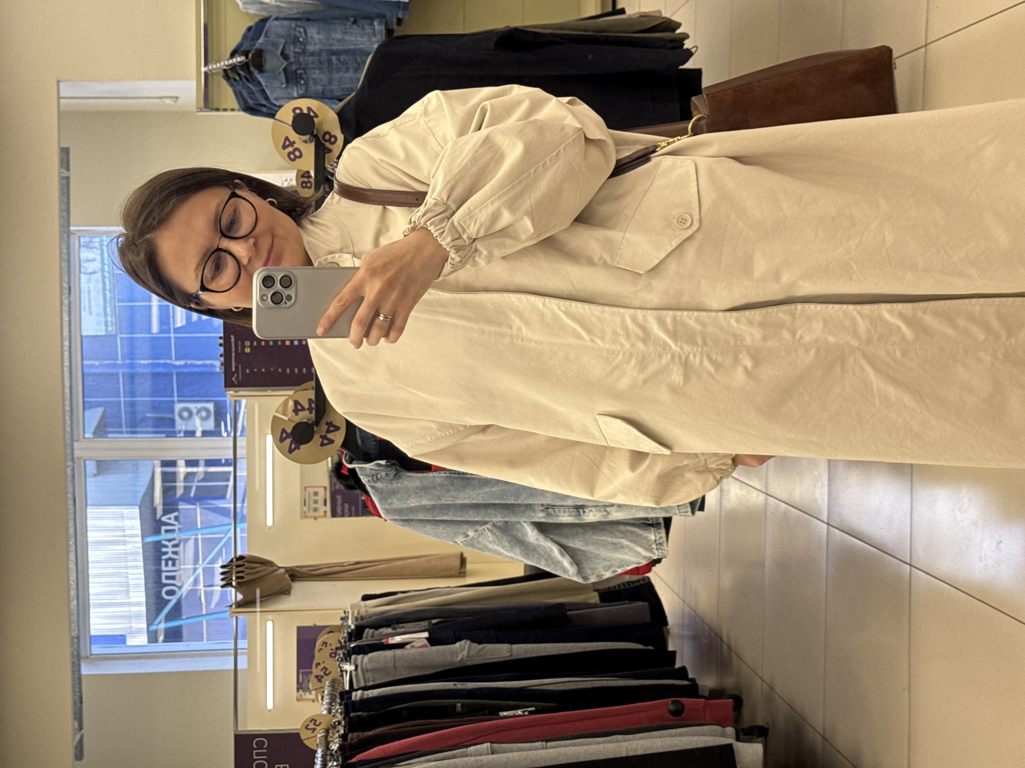 A woman with eyeglasses is taking a selfie in a clothing store. She is holding a gray iPhone and wearing a long, beige trench coat with a brown leather shoulder bag strap visible. The store has clothing racks filled with items including jeans, trousers, and other garments. A window with a reflection of a building with the word "ОДЕЖДА" (clothing) in Russian is visible. There are clothing size labels hanging from the racks. The location is likely a clothing store in Izhevsk, Russia, given the Russian text on the window.