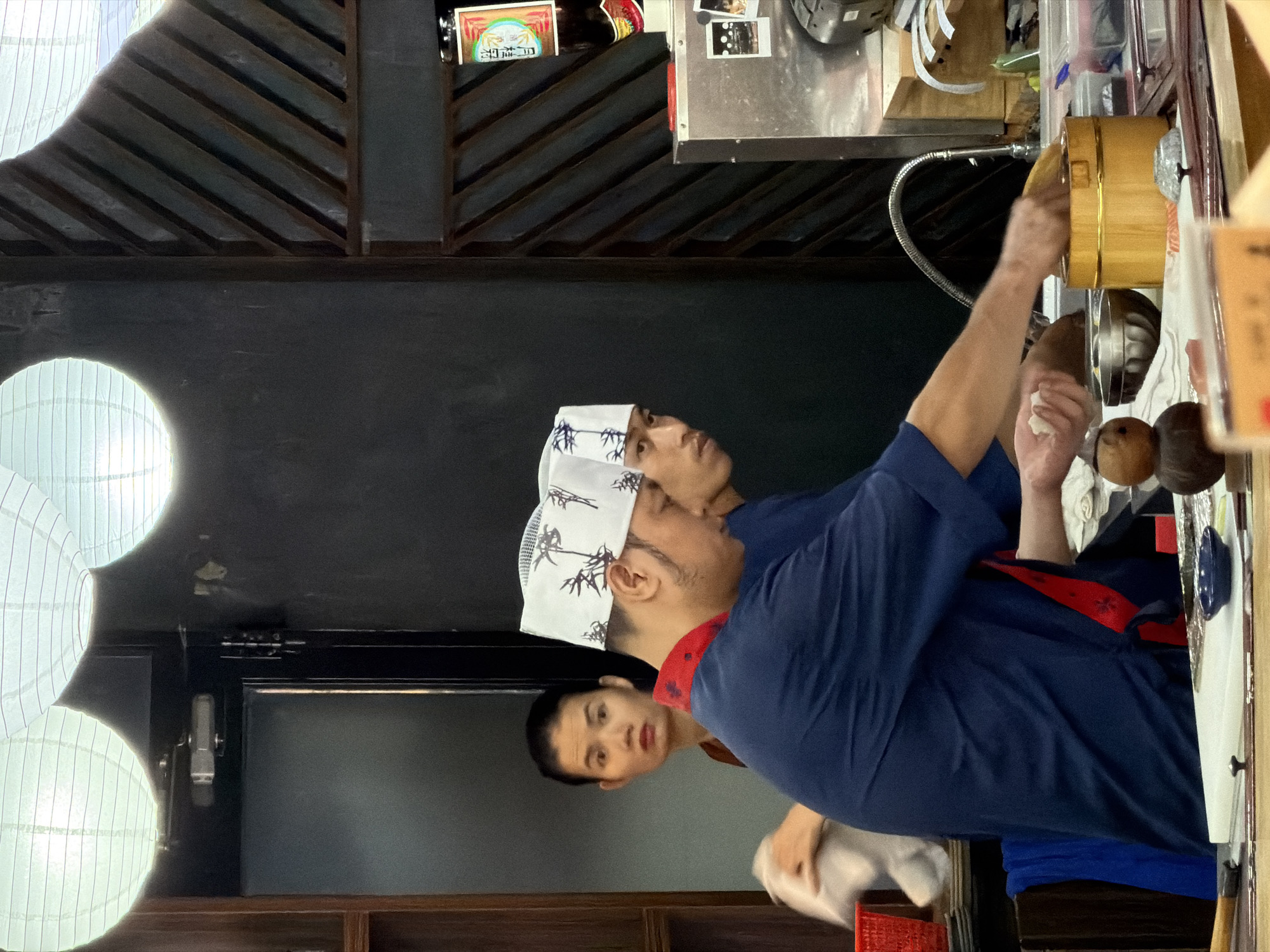 In a dimly lit sushi restaurant, possibly in Nha Trang Ward, Vietnam, two sushi chefs are preparing food behind a counter. The chef in the foreground, wearing a blue uniform and a white cap with a bamboo pattern, is focused on his task, his right hand holding a piece of rice. To his left, another chef, also in a blue uniform and white cap, is partially visible, looking down. Behind them, a woman with short black hair and wearing a brown garment stands near a doorway, holding a white cloth. The counter is cluttered with various items, including bowls, a wooden container, and what appears to be raw fish. The restaurant's ambiance is further suggested by the presence of traditional Japanese lanterns hanging from the ceiling and wooden partitions on the wall.