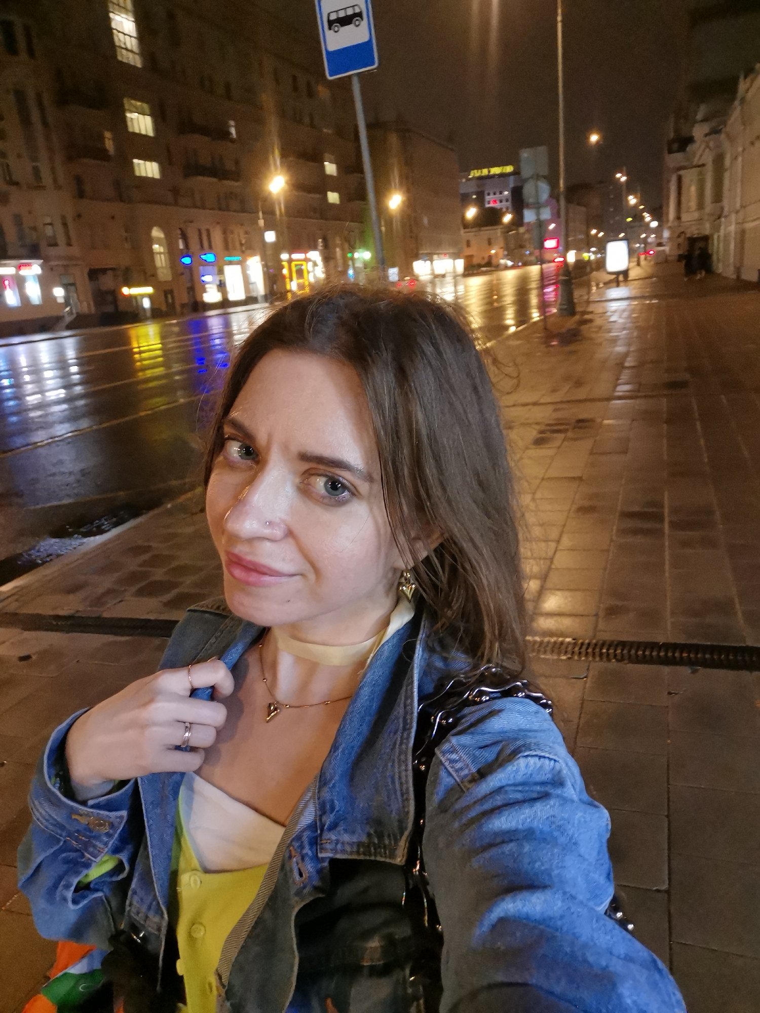 A woman with long brown hair is posing for a selfie on a city street at night. The street appears wet, reflecting the lights from the surrounding buildings. She is wearing a denim jacket over a light-colored top and a necklace. A bus stop sign is visible in the background. The setting is a city street at night with illuminated buildings and wet pavement. The city is Unknown, Unknown.
