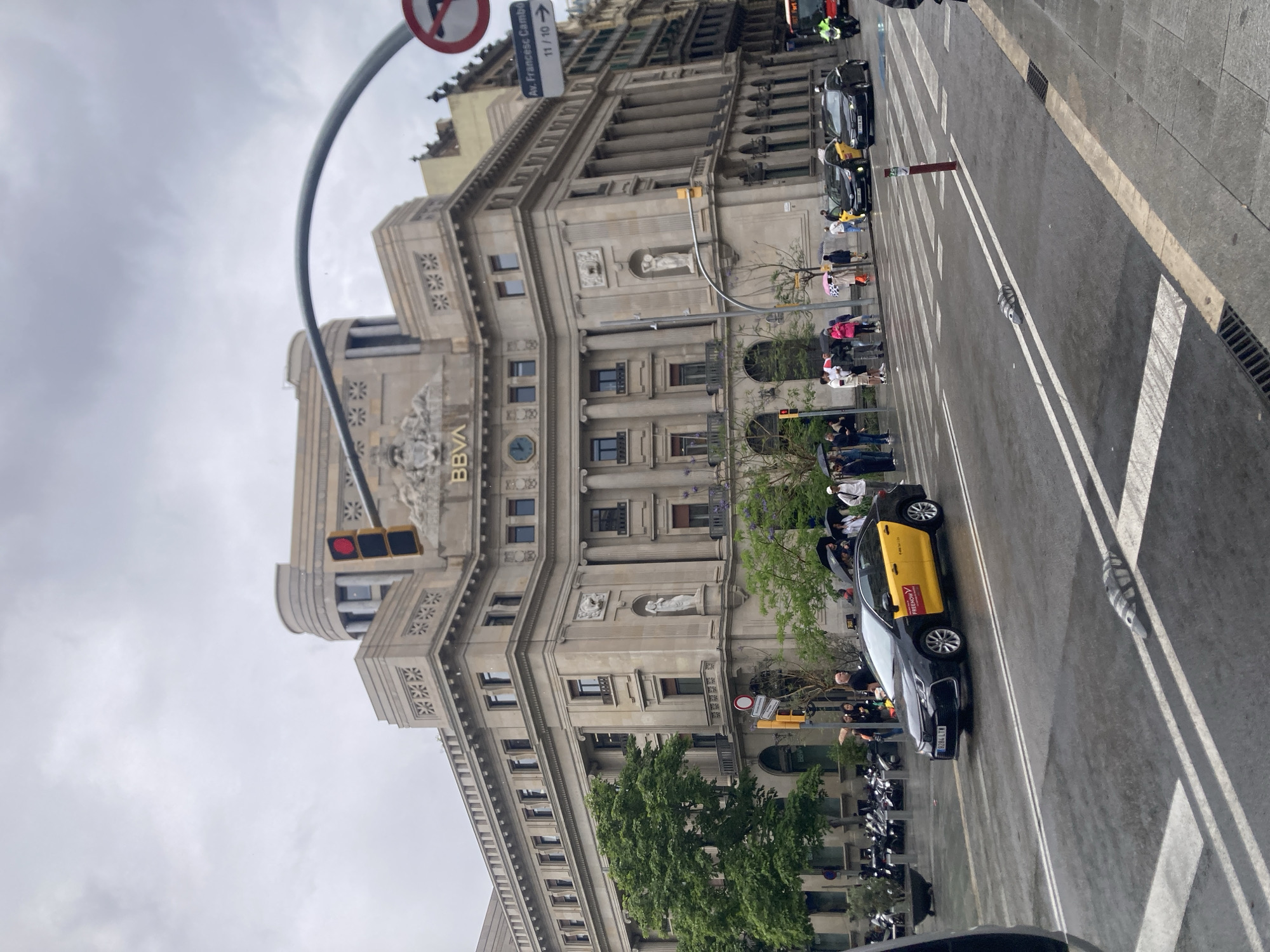 The image depicts a street scene in Barcelona, Spain, dominated by a large, ornate building identified by the "BBVA" signage. The building has a classical architectural style with columns, statues, and decorative elements. Traffic lights are visible on the building's facade.
On the street level, a line of taxis, with their distinctive yellow doors, is visible along the street. Several pedestrians are grouped on the sidewalk waiting to cross at a crosswalk. In the background, a street sign indicates the direction towards Av. Francesc Cambó. A no-pedestrian sign hangs over the sidewalk. The weather appears overcast.