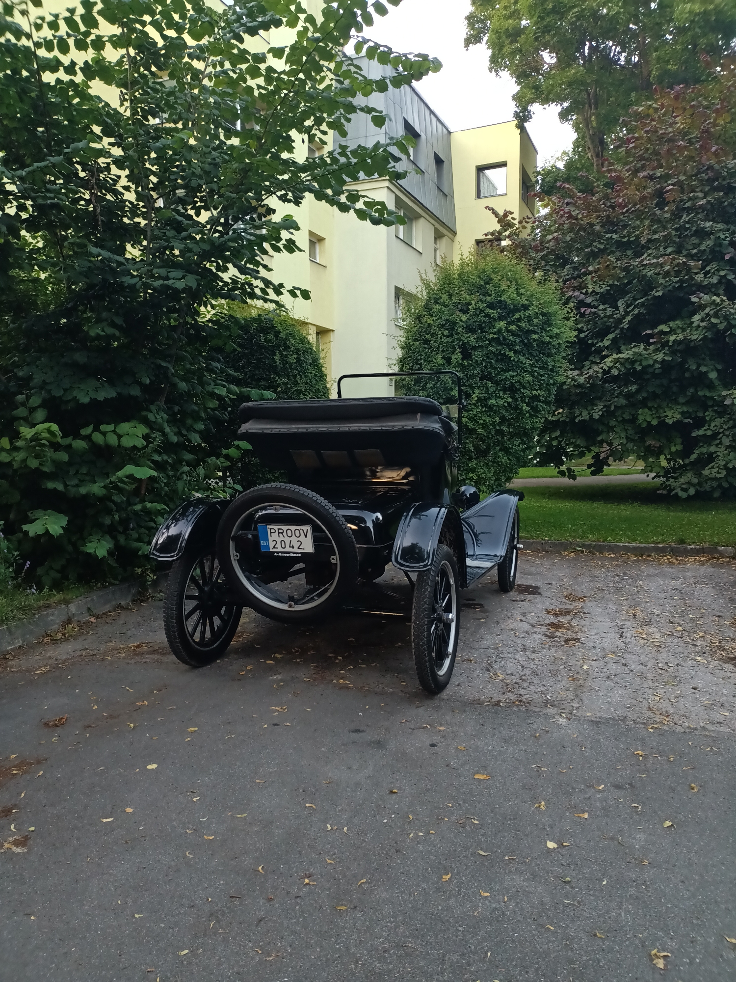 The image depicts the rear view of a black vintage car parked on a paved area with scattered fallen leaves. The car has a spare tire mounted on the rear. A license plate is visible on the spare tire, reading "PROOV 2042 EST A-Alberte.ee".
In the background, there is a modern building with light yellow and gray facades, featuring several windows. Lush green trees and bushes surround the building and the parking area. The scene suggests a residential or public space within a city.
Based on the license plate, the location is identified as Tallinn, Estonia.