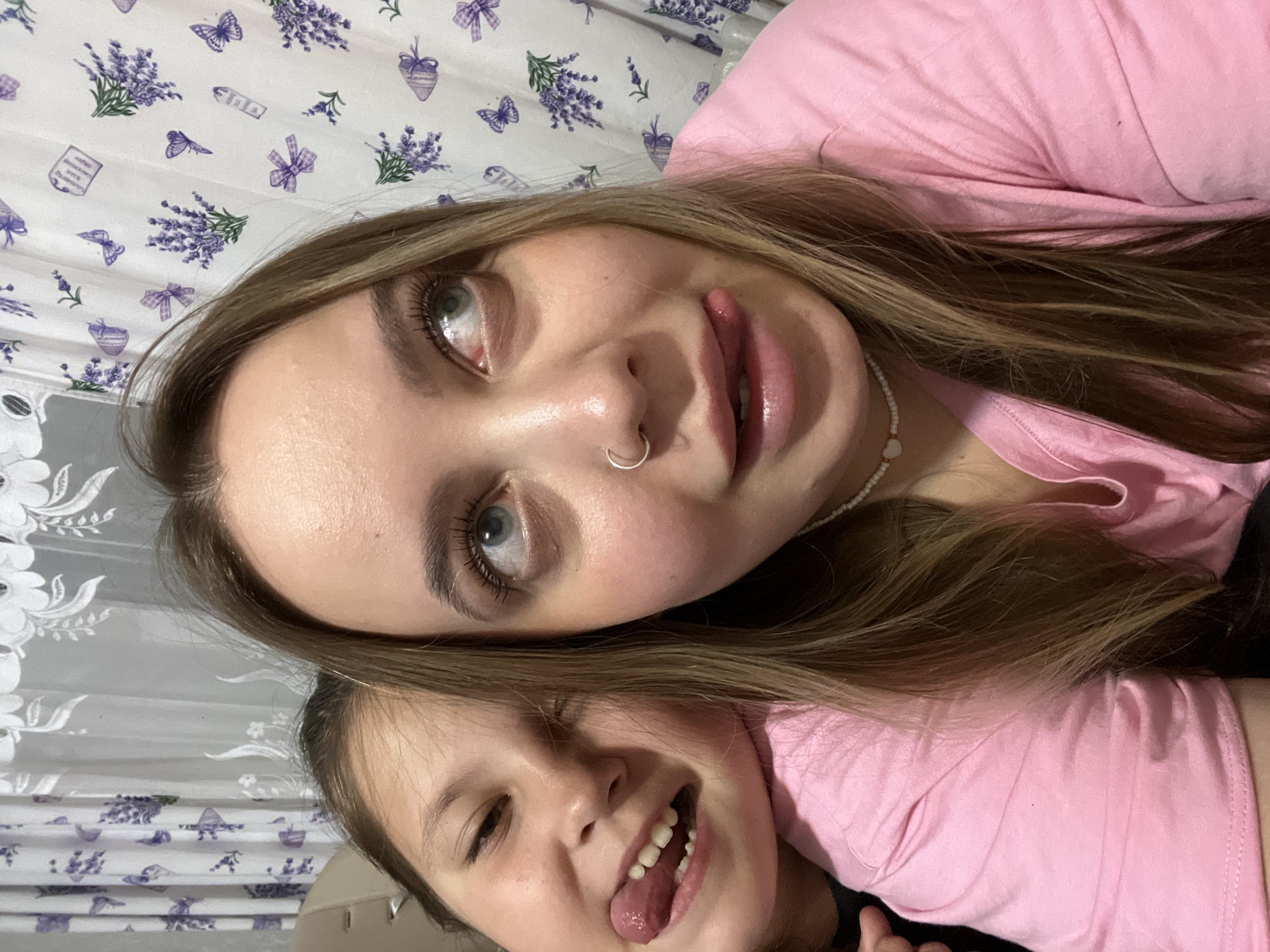 A close-up, angled shot captures two females, an adult and a child, in what appears to be an affectionate and playful moment indoors. Both subjects wear pink tops and have light brown hair.
The adult, positioned towards the top of the frame, has blue-grey eyes, a nose ring, and a white beaded necklace. Her long hair cascades over her shoulder, and she looks upwards with her tongue slightly extended. The child, nestled below the adult, smiles widely, her tongue also extended, revealing missing front teeth. She looks directly towards the viewer or camera.
The background consists of a white fabric patterned with various purple motifs, including lavender sprigs, butterflies, bows, and small rectangular shapes that might contain faint, illegible text. This pattern suggests a casual home environment, possibly bedding or a curtain.
The overall emotion conveyed is one of joy and playfulness, indicative of a warm familial bond. The time of day is not discernible due to the indoor setting. The location context provided is Novopokrovskaya, Russia.