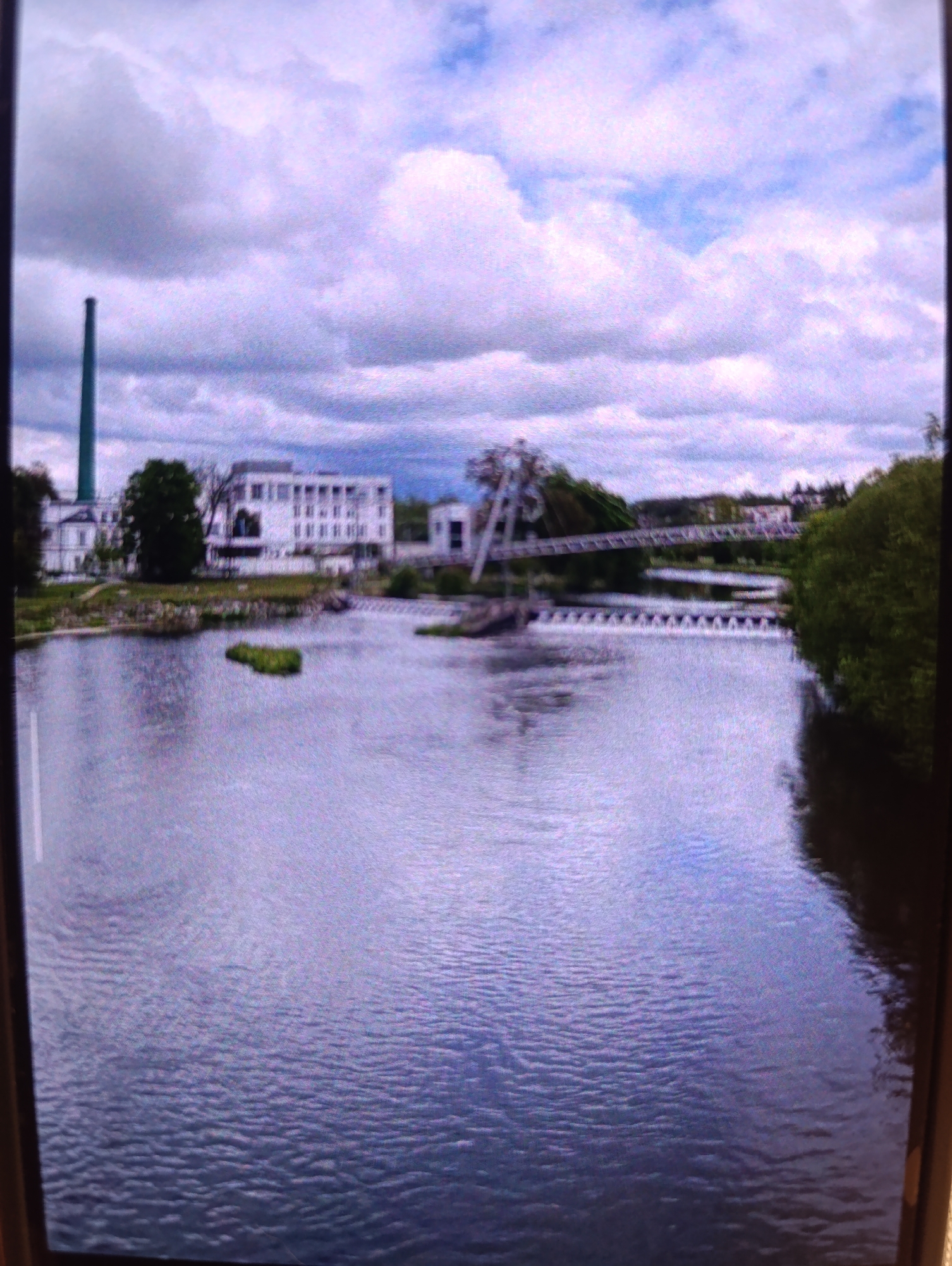 The image shows a wide body of water in the foreground, with ripples on its surface. In the mid-ground, a dam with a series of small cascades is visible across the water. Beyond the dam, a tree-lined bank separates the water from a cityscape. Several buildings are present, including a large, modern white building with multiple floors and windows, and a tall, slender green smokestack. A bridge with an unusual, angular structure is seen spanning the water in the distance, with a line of trees and more buildings on the far bank. The sky above is filled with dramatic clouds, some appearing white and fluffy, others darker and more ominous, with patches of blue sky visible between them. The overall impression is that of an industrial or post-industrial area situated alongside a river.
