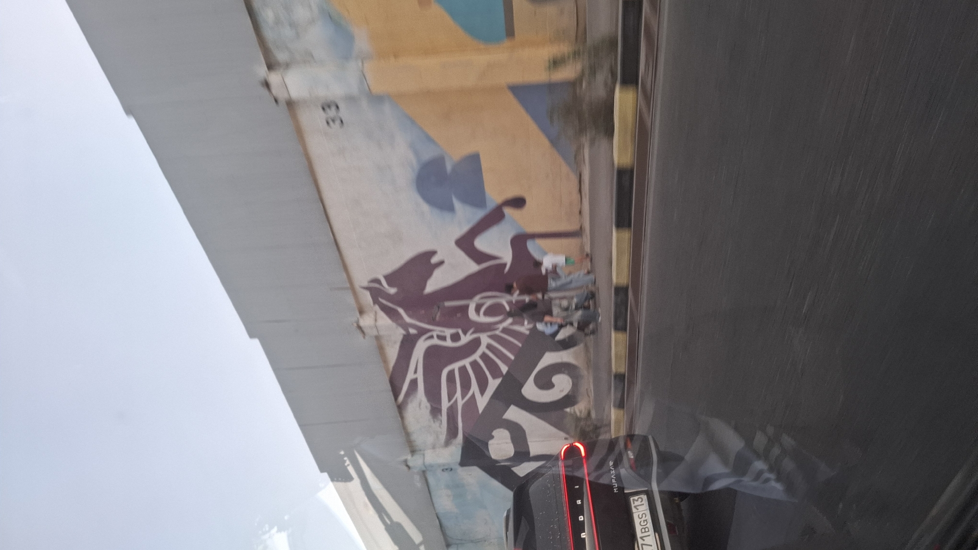 The image shows a mural of a winged horse on a concrete wall in Shymkent, Kazakhstan. The mural is predominantly purple and white, with some blue and yellow geometric shapes integrated into the background. In front of the wall, a dark-colored car, identified by its license plate as 71 BGS 13, is parked or driving on a paved road. The car has red taillights illuminated. Three people are visible standing near the wall, two adults and one child. The environment appears to be outdoors, with a clear sky. The wall itself has a lighter colored structure above it, and a sidewalk with yellow and black bollards separates the wall from the road. The overall scene is urban.