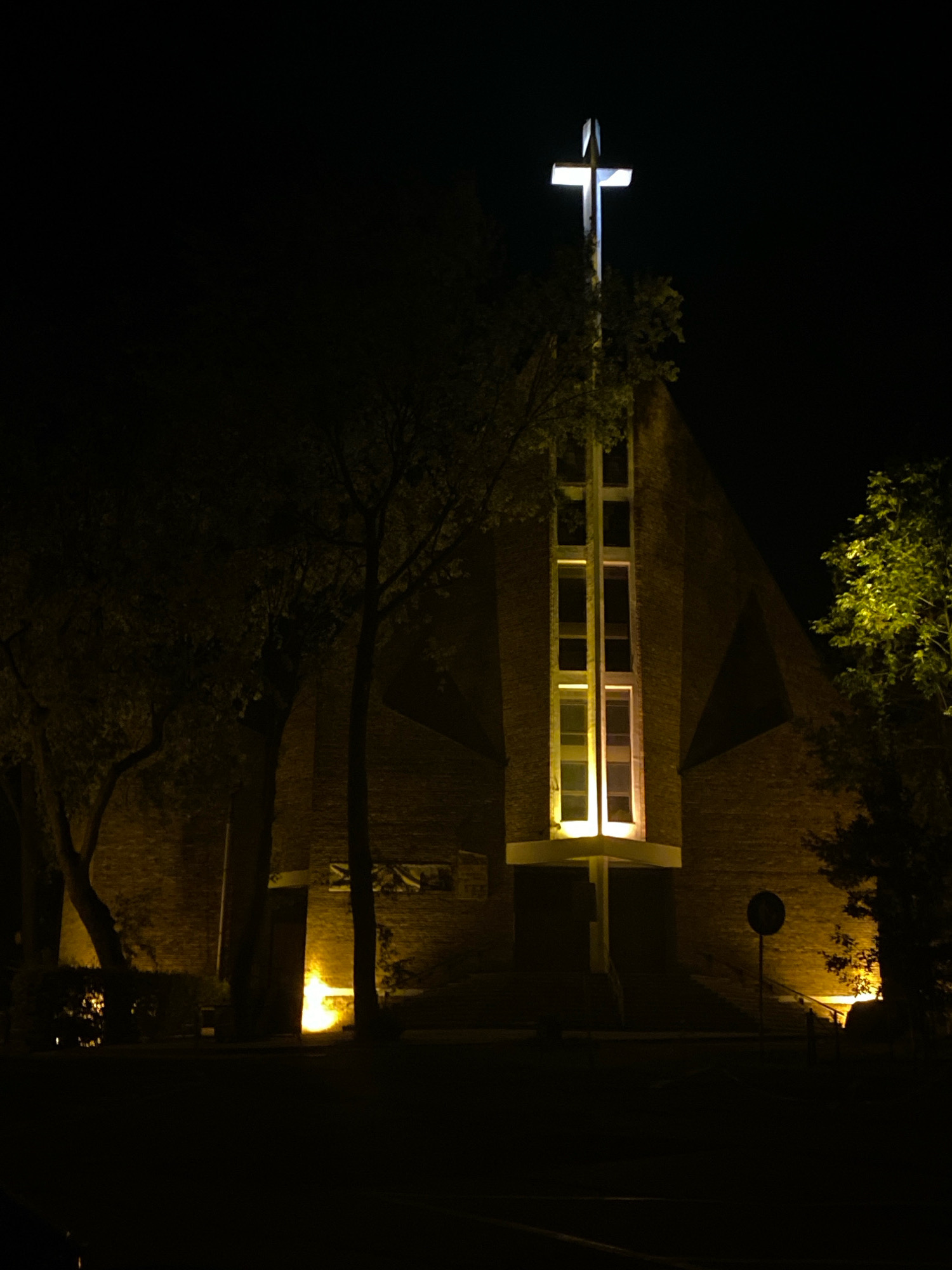 The image depicts a church building at night, illuminated by its external lights. A prominent cross, glowing brightly, is positioned on top of the structure. The church is constructed from brick and features a tall, narrow window with multiple panes, also lit from within. Trees surround the building, their foliage visible against the dark sky. The foreground is obscured by darkness, with only hints of ground cover and a street sign visible. The scene suggests a quiet, possibly solemn, atmosphere. The specific location of this church in Katowice, Poland, is not identifiable from the visual information provided in the image.