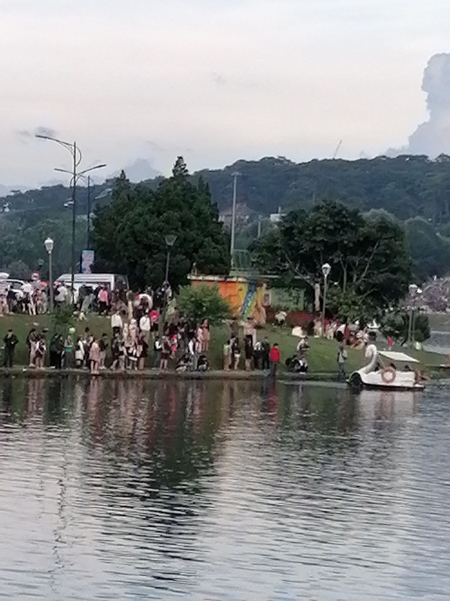 The image shows a cloudy day scene by Xuan Huong Lake in Da Lat, Vietnam. A crowd of people is gathered on the grassy embankment, some standing and others sitting. Several cars and a white van are parked on a paved area behind the crowd. In the lake, a small white boat with a canopy is visible, with at least one person in it. The background features lush green trees and hills under a hazy sky, with some buildings and structures faintly visible. The overall atmosphere appears to be that of a casual gathering or recreational activity.