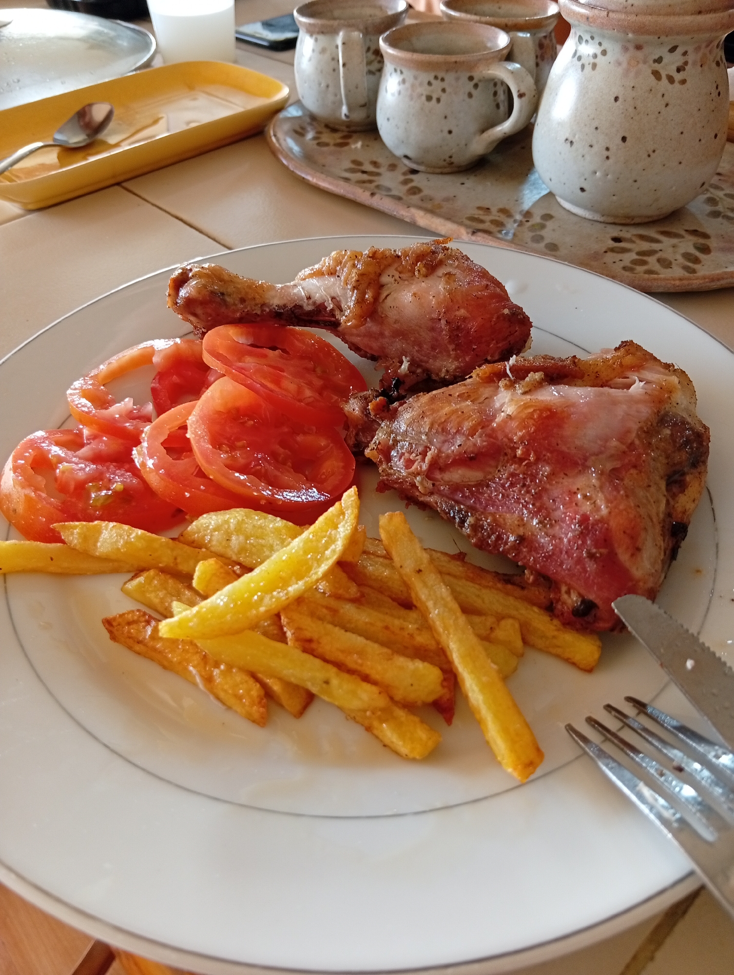 A close-up overhead shot shows a white plate with a meal of fried chicken, sliced tomatoes, and french fries on a light-colored tiled surface. The chicken appears to be roasted and browned, with visible skin and meat. The tomatoes are sliced into rings and arranged around the chicken. A pile of golden-brown french fries is situated at the bottom of the plate. A fork and knife are visible at the bottom right of the plate, with the knife partially obscuring the fries. In the background, out of focus, are several ceramic mugs and a jug with a speckled pattern, resting on a decorative tray. A yellow dish with a spoon is to the left of the plate. The overall scene suggests a casual meal setting. The presence of the specific decorative ceramic set and the style of the meal implies it is likely within a residential or restaurant setting in San Antonio de Los Altos, Venezuela.