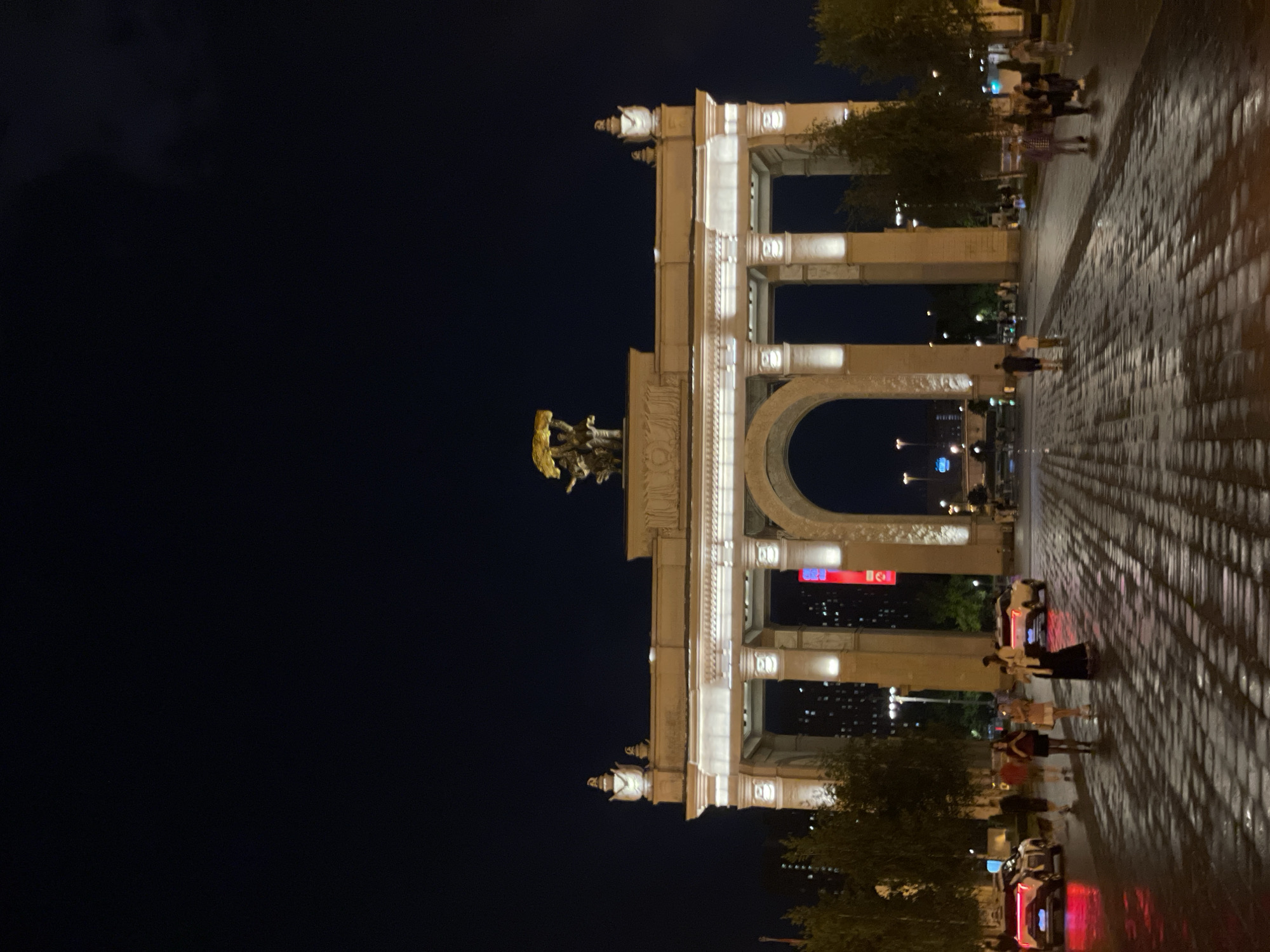 The image depicts the Triumph Arch in Moscow, Russia, at night. The arch is brightly lit and its neoclassical architectural details are clearly visible. A statue atop the arch is also illuminated. Pedestrians are walking on a paved surface in front of the arch. Some cars are also visible. The sky is dark. The scene is viewed from a street-level perspective.