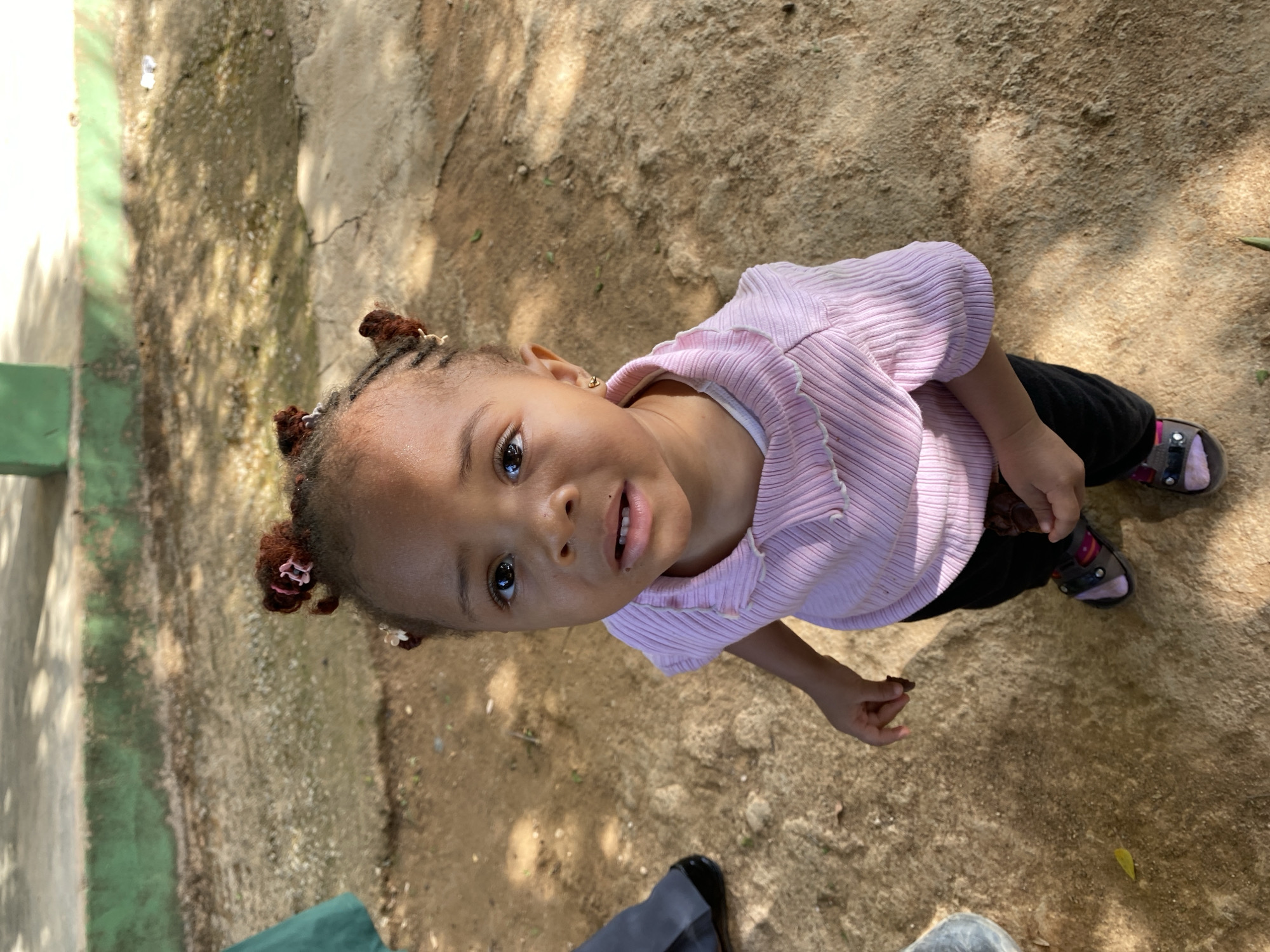 A young child stands on a dirt and concrete ground surface, likely outdoors. The child wears a light pink, ribbed shirt, dark pants, and sandals. Their hair is styled in braids. The environment appears to be a shaded area, with possible foliage present, based on the shadow patterns. The location is Gura Top, Nigeria. An adult's legs and feet are visible in the lower portion of the image. The child is looking up towards the camera.