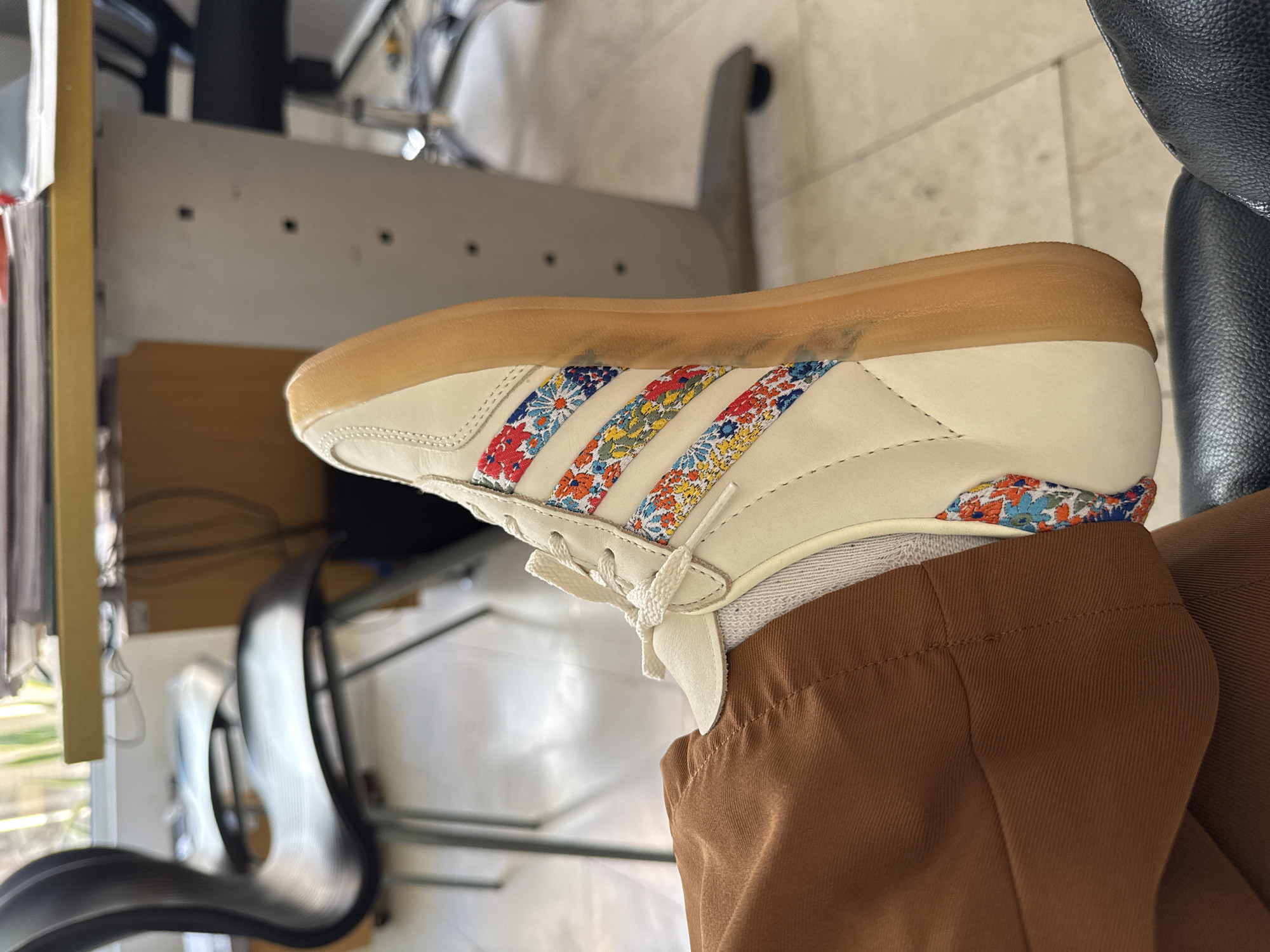 A close-up shot shows a person wearing a white sneaker with floral stripes. The sneaker has a gum sole and is paired with brown pants. The setting appears to be indoors, with office furniture and a chair visible in the background. The image does not provide enough information to determine the location in Pampatar, Venezuela.