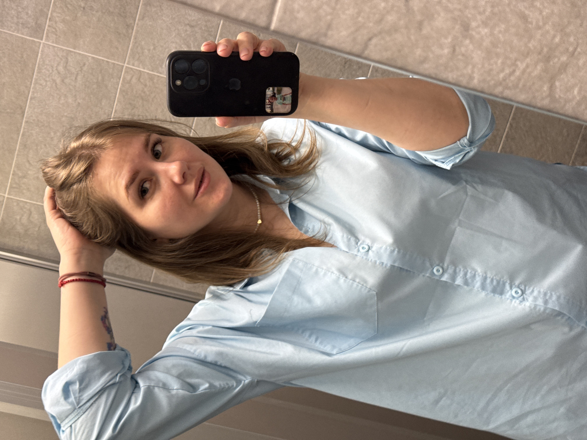 A woman is taking a selfie in a bathroom with tiled walls. She is holding a black smartphone in her right hand, partially obscuring her face. She is wearing a light blue button-down shirt and a pearl necklace. Her left hand is touching her hair. There are two thin red bracelets on her left wrist and a visible tattoo on her left forearm. The location is in Perm, Russia, as the reflection in the phone reveals a mirror and a person inside the restroom.