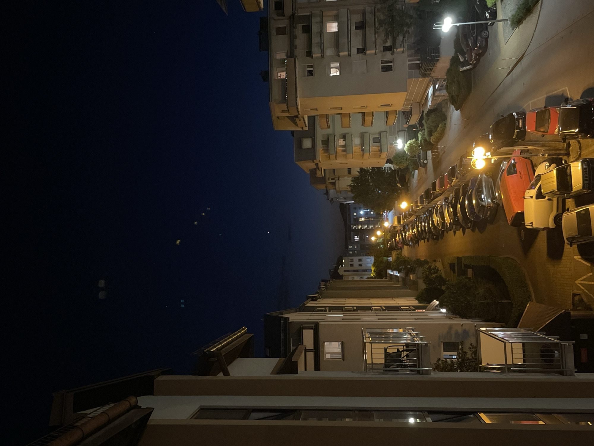View from a balcony in Unknown, Unknown, at nighttime, facing parallel buildings on a street. There are several parked cars and lampposts on the right and the sky on the left.