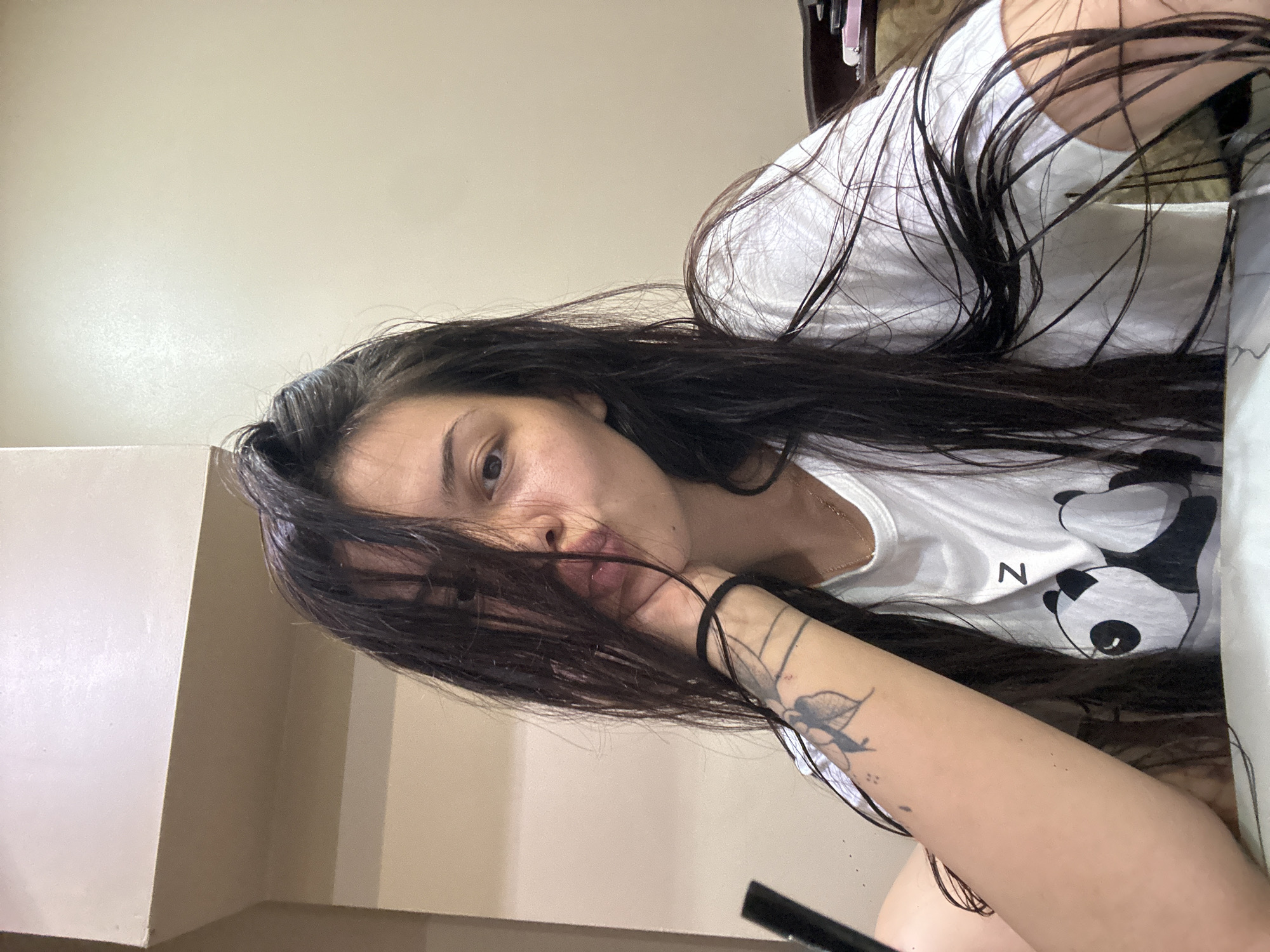 A young woman with long, dark, wet hair is leaning her head on her hand, with her mouth slightly pursed. She is wearing a white t-shirt with a panda design and the letter 'Z'. She has a tattoo on her left forearm. The background is a plain, light-colored wall. The scene appears to be indoors. There is no indication of the location being in Sliven, Bulgaria.