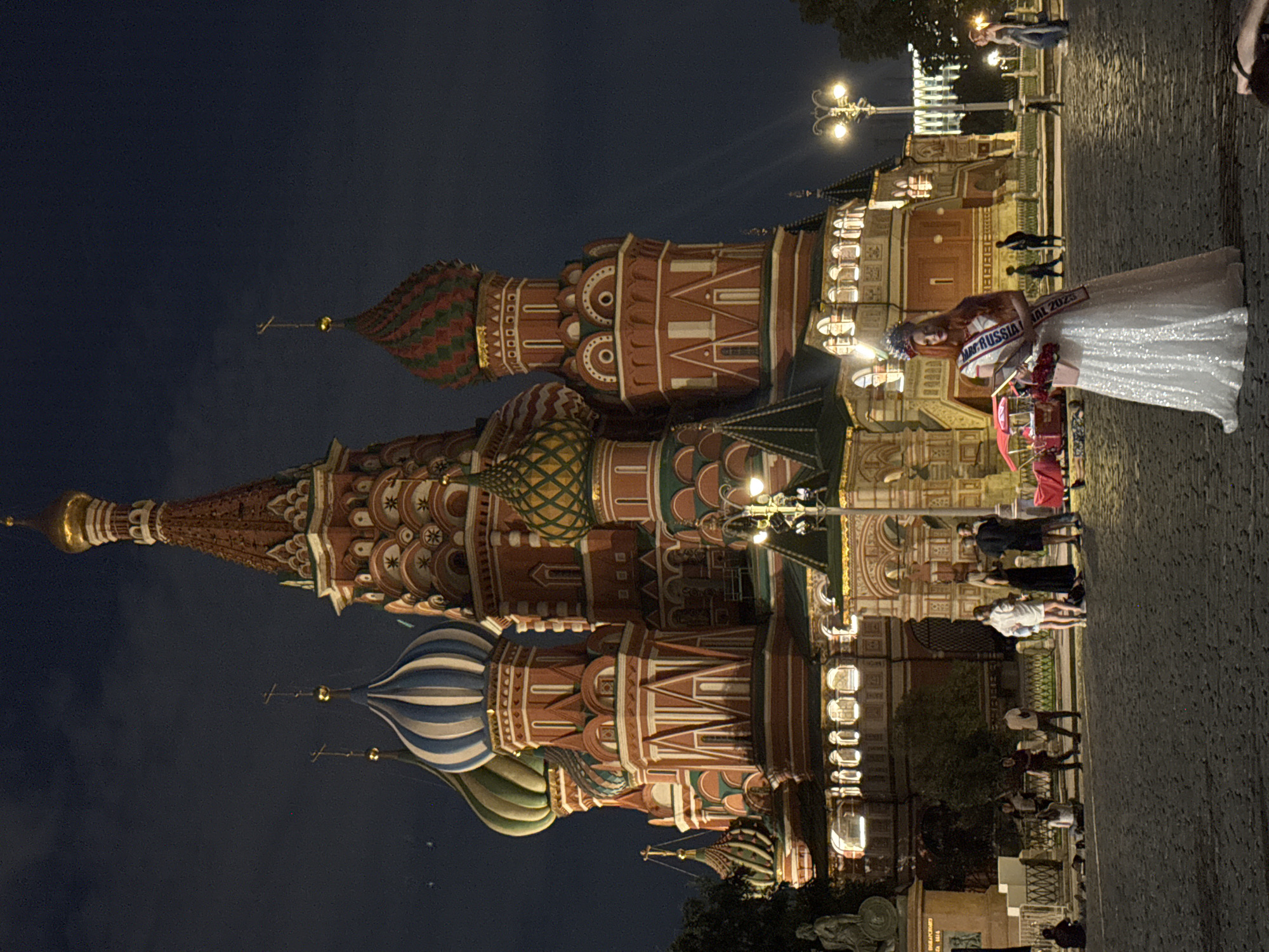 The image shows a night view of St. Basil's Cathedral in Moscow, Russia, with its iconic brightly colored onion domes illuminated. In the foreground, a woman is standing, dressed in a white sequined gown and wearing a sash that says "MR RUSSIA NATIONAL 2023". She is also wearing a crown and holding a bouquet of red flowers. She appears to be posing for a photograph. Other people are visible in the background, some are standing and talking, while others are sitting on the cobblestone ground. The sky is dark, indicating it is nighttime, and streetlights are illuminating the scene.
