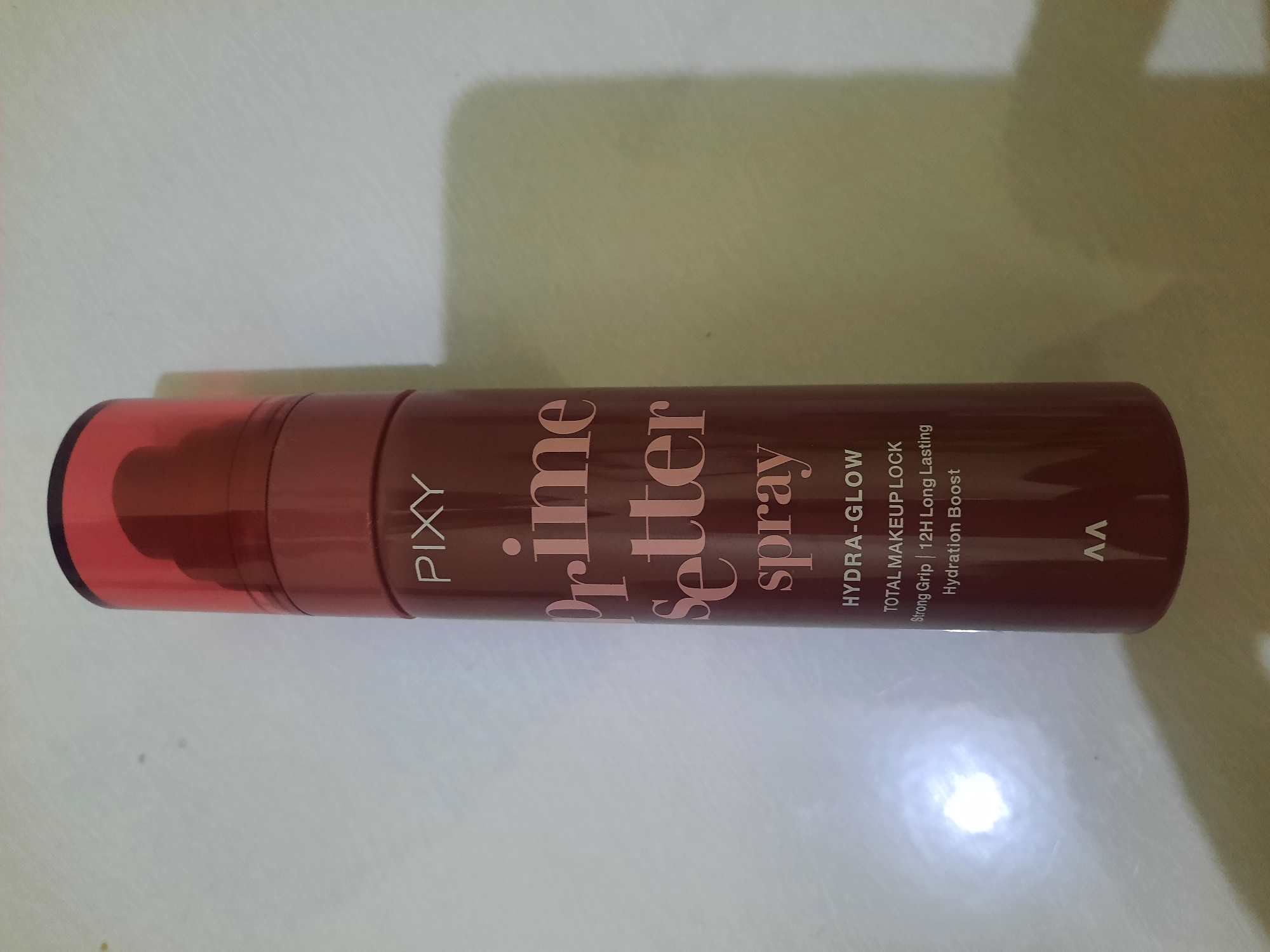The image displays a tall, dark red spray bottle of Pixy Prime Setter Spray, with pink lettering. The bottle is upright and centered in the frame, against a plain, light-colored background that appears to be a surface. The lighting in the image creates a slight shadow to the right of the bottle and a bright, circular reflection on the surface to the left. The location in Lhokseumawe, Indonesia, is not discernible from the visual information in the image itself.