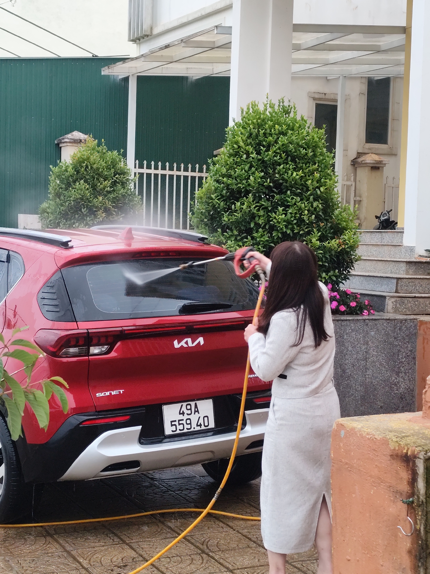 A person is washing a red Kia Sonet car with a pressure washer in Lạc Dương, Vietnam. The car is parked on a tiled driveway in front of a house with white pillars and steps. The person, wearing a light-colored outfit, is holding the pressure washer wand and aiming it at the rear windshield of the car. The driveway and car are wet, indicating that the washing is in progress. Lush green bushes and a white fence are visible in the background, along with a green corrugated metal wall. A motorcycle is parked on the steps of the house. The license plate of the car reads "49A 559.40".