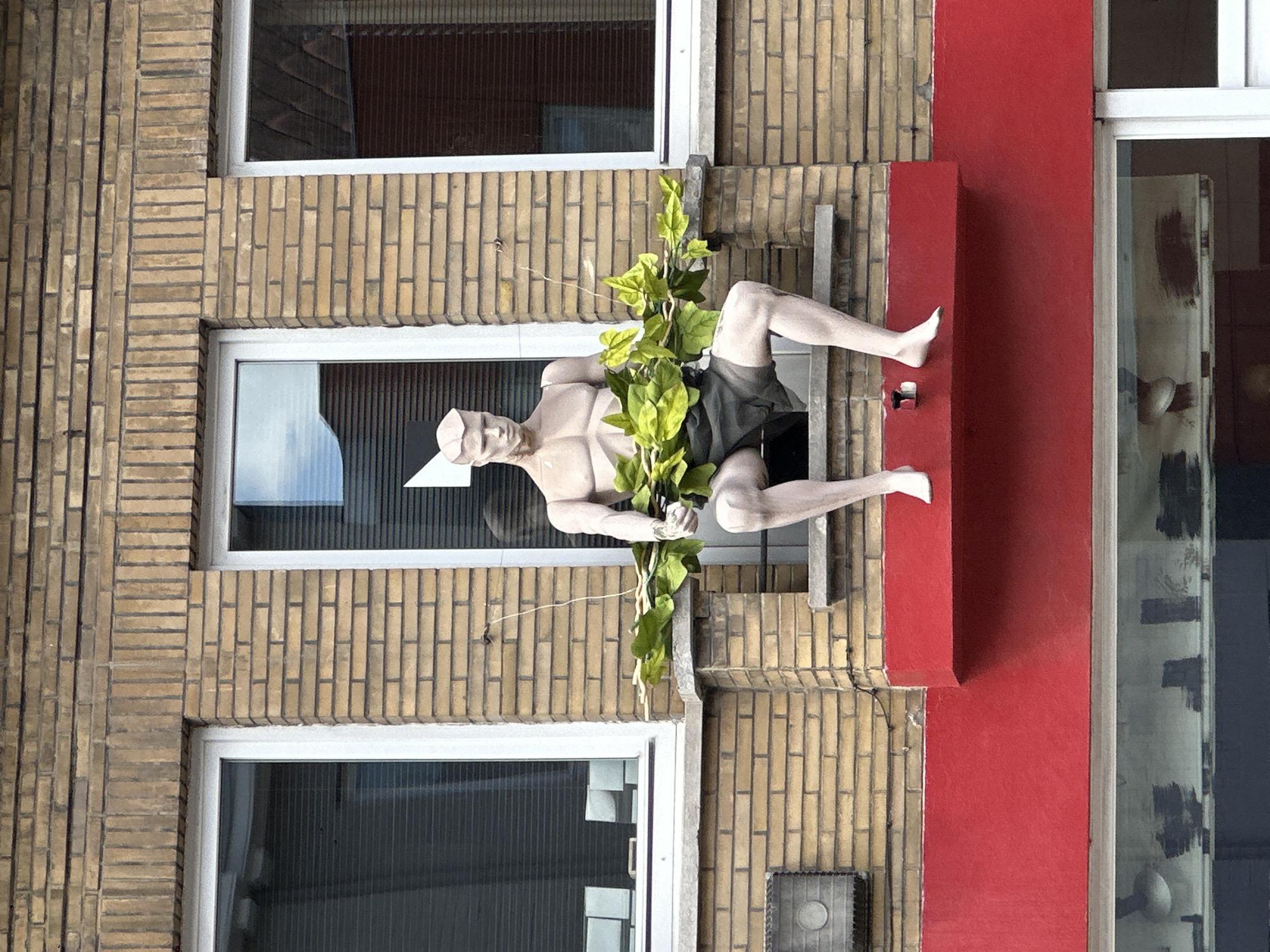 The image shows a male mannequin with a muscular physique sitting on a ledge outside a building. The mannequin is wearing dark shorts and holding a branch with green leaves. The building has a brick facade and white window frames. A bright red ledge is visible below the mannequin. The scene appears to be in an urban environment, possibly Bruges, Belgium, based on the architectural style.