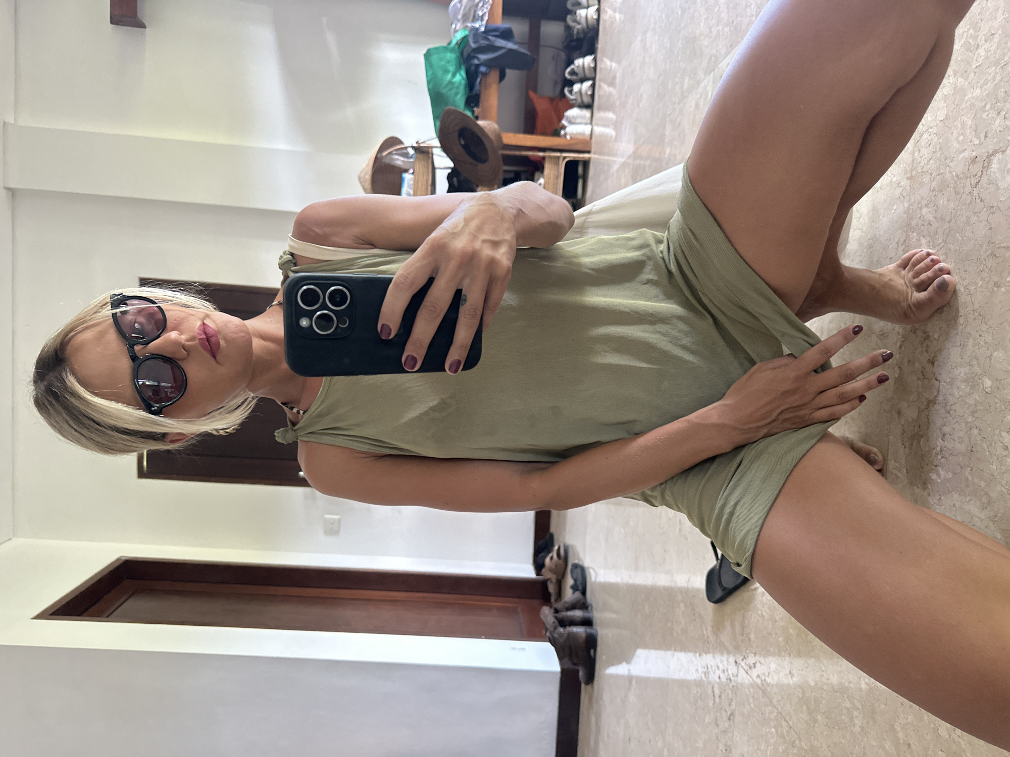 A woman takes a selfie indoors. She wears a light green dress and sunglasses, and holds a black smartphone. The location appears to be a house in Kuta Selatan, Indonesia, judging by the tropical decor and light. The environment includes doors, a closet with items like shoes and bags, and a marble floor.