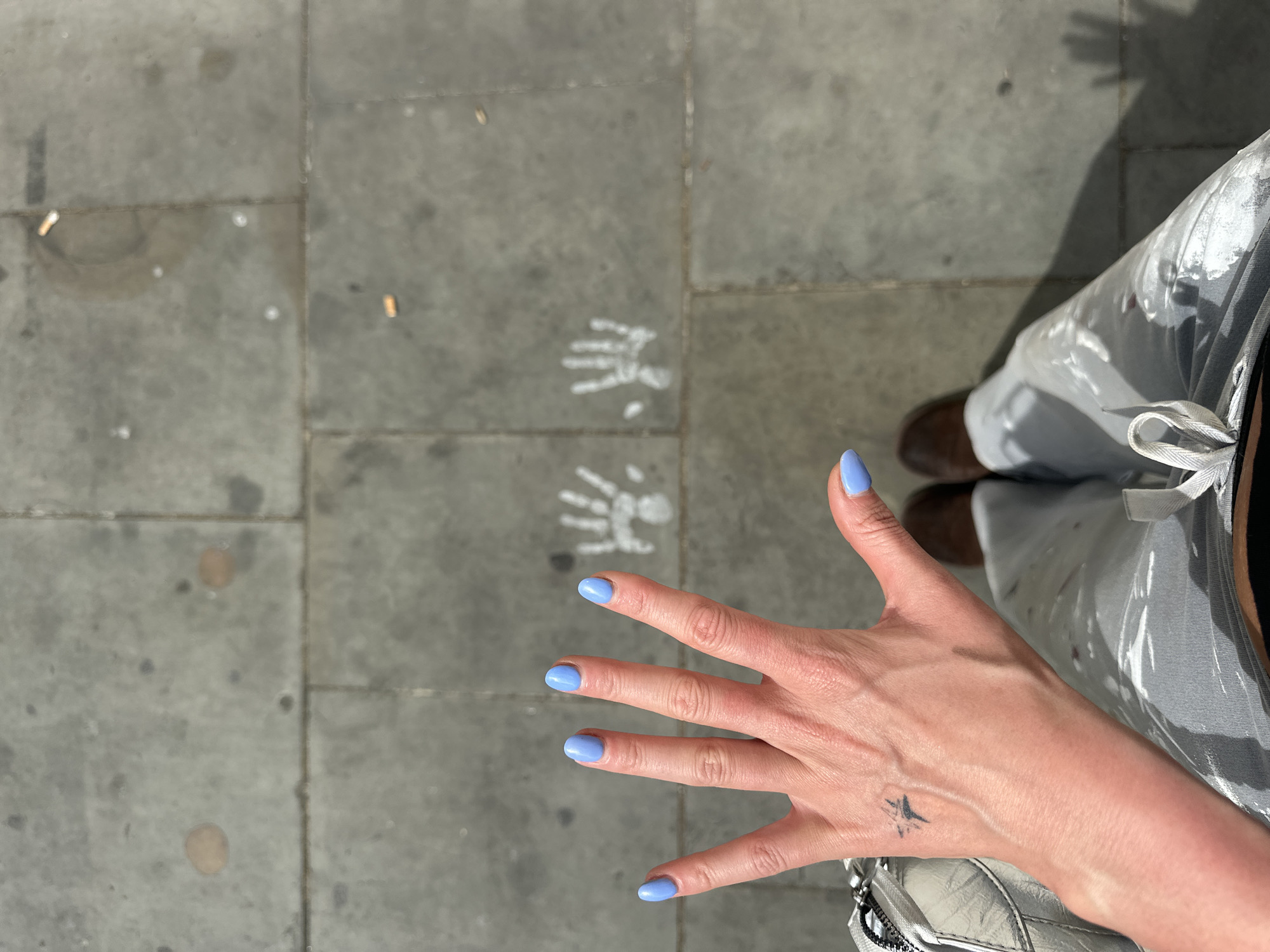 A top-down shot presents a person standing on a gray paved street in London, United Kingdom, adjacent to two chalk handprints on the pavement. The person's left hand is outstretched, fingers spread, revealing light blue nail polish and a small star tattoo on the wrist. The individual is wearing light gray pants and brown shoes. Cigarette butts are scattered on the pavement.