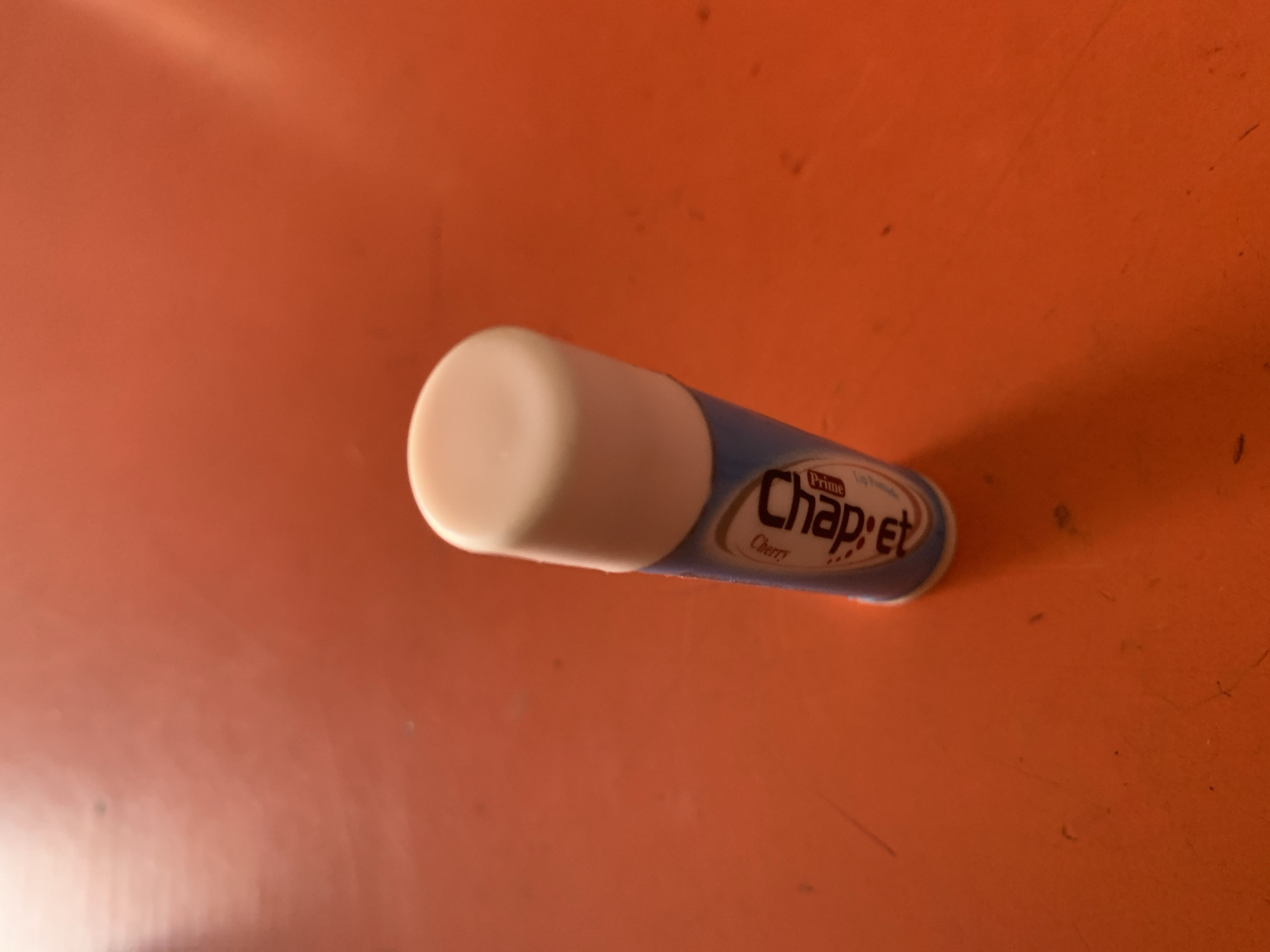A close-up, medium shot shows a single tube of Chapstick lip balm standing upright on an orange, textured surface. The lip balm tube is predominantly blue with a white cap and a white label that features the "Chapstick" logo in large black letters, with "Cherry" written above it in smaller, curved white text. The "Prime" logo is visible in red on the lower right side of the label. The surface appears to be a table or countertop, with some subtle dust or debris visible upon it. The lighting creates a shadow to the left of the lip balm and a highlight on the upper right portion of the tube. The background is a blurry expanse of the same orange surface. There are no people or animals present in the image.