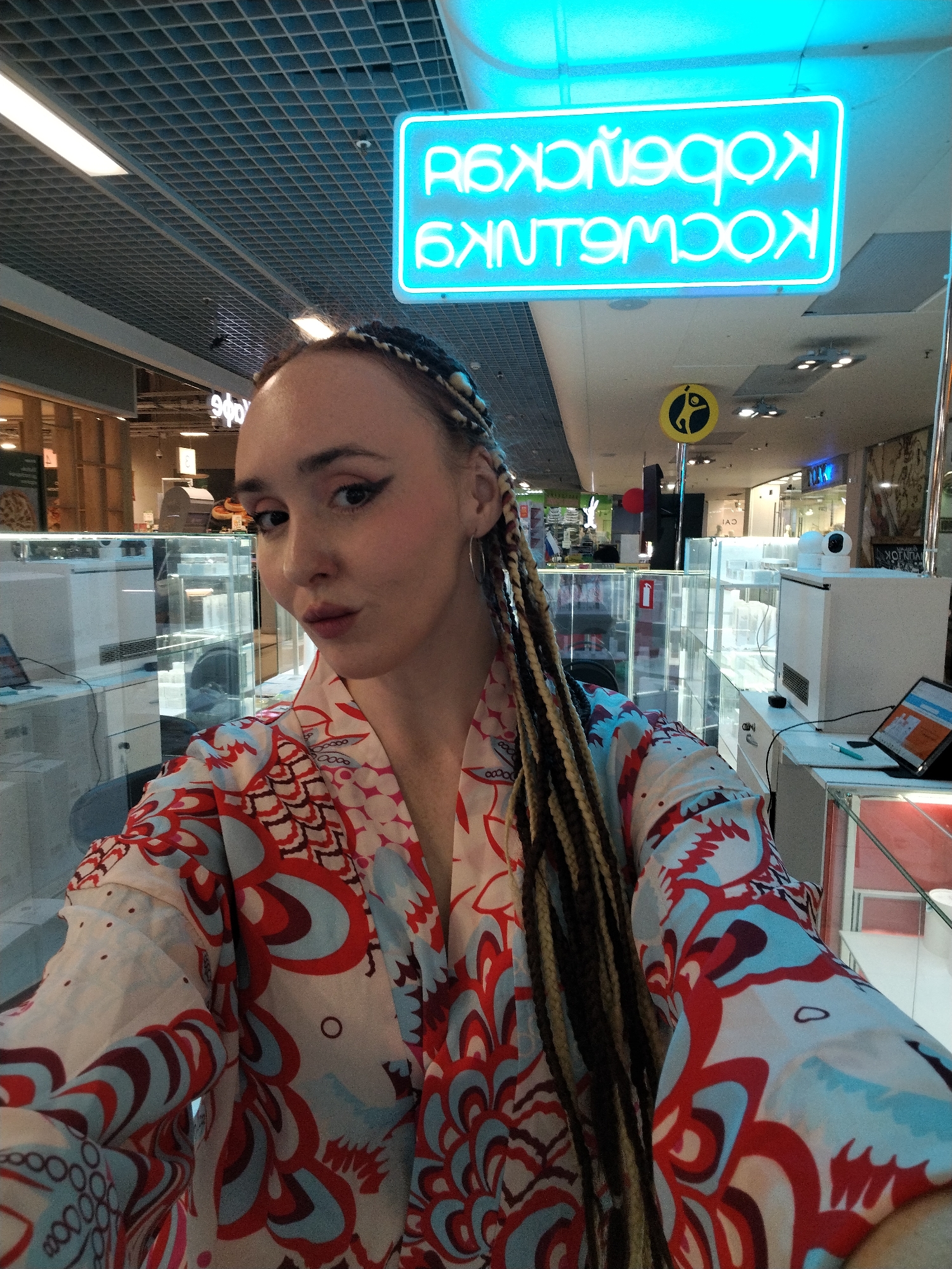 The photograph is a selfie of a light-skinned woman with blonde and brown braided hair and a pursed mouth. She wears large hoop earrings and a colorful patterned shirt with swirling red, white, and blue designs.
Behind her is the interior of a commercial space, possibly a shopping mall or market. There are glass display cases, some with screens or devices on top, indicating a retail environment. A bright neon sign with Cyrillic characters is visible, indicating the city of Izhevsk, Russia.
The background features a grid-patterned ceiling and other storefronts, suggesting a bustling commercial area. A yellow sign with a figure is also visible. The overall setting implies a public space within a retail establishment.