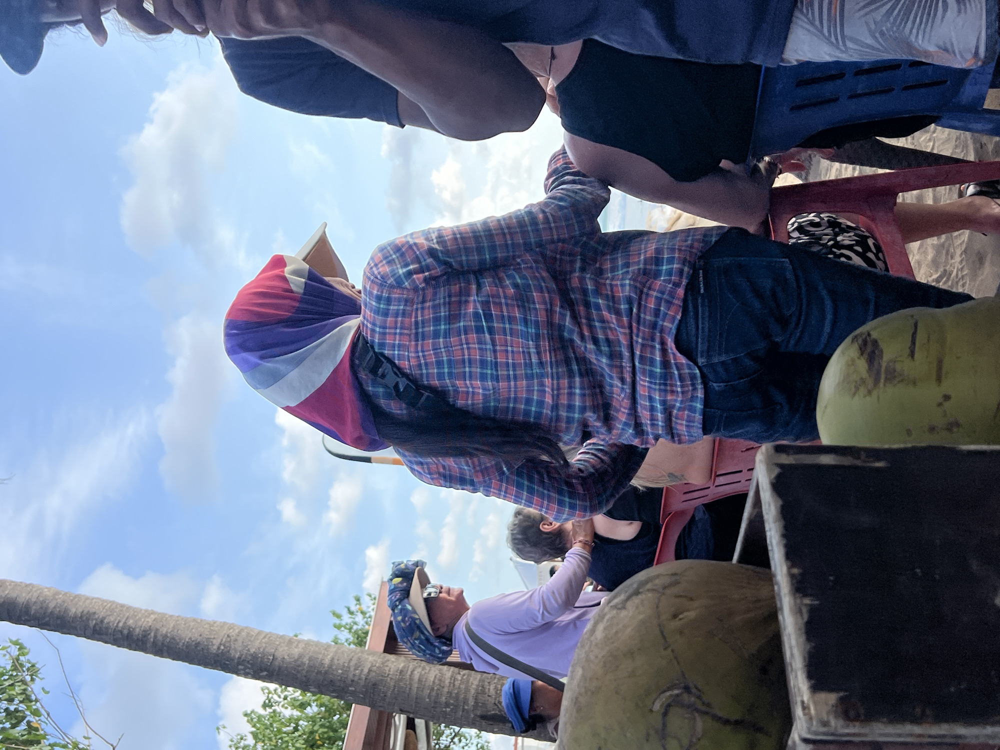 In Kuta, Indonesia, on a beach with sandy ground and a partly cloudy sky, a group of people are gathered. A woman in the foreground, facing away from the camera, wears a colorful headscarf and a plaid shirt over dark jeans. To her left, another woman is seated, wearing a light purple hoodie and a wide-brimmed hat. To the right, a person in a dark t-shirt is visible from the shoulders down, and another individual sits in a blue chair. In the lower left corner, two coconuts are present, one in a black container. A palm tree trunk stands on the left side of the image.