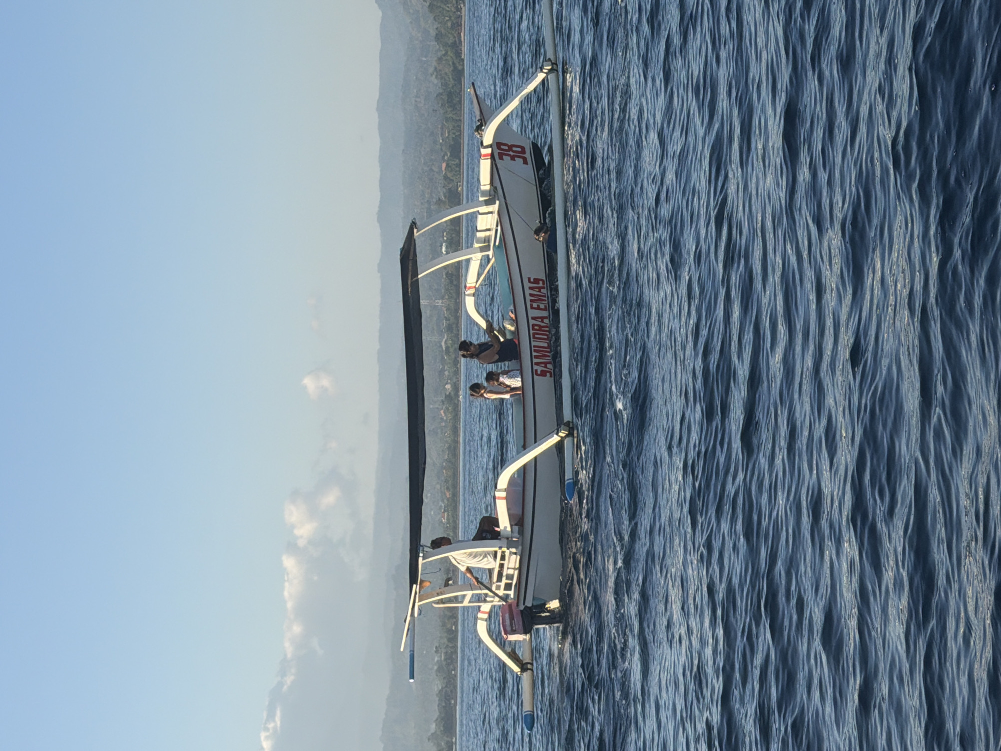 A traditional outrigger boat, identified with the name "SAMUDRA EMAS" and the number "38," travels across a body of water. The boat has a dark canopy providing shade and is powered by an outboard motor. Several people are on board: a man is steering the boat with an oar, and two women and two children are seated behind him. In the water near the boat, one person is visible, possibly swimming or snorkeling. The background shows a coastline with lush green hills and a hazy sky. The scene appears to be set in a tropical or subtropical coastal region.
