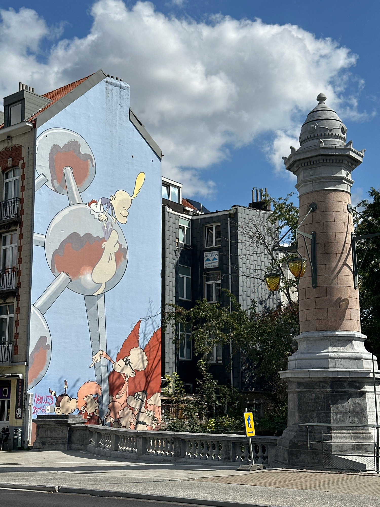 The image shows a mural on the side of a building in Brussels, Belgium, depicting characters from "The Adventures of Tintin" in front of the Atomium. The mural features a light blue background with a large illustration of the Atomium structure. Several Tintin characters, including Tintin himself, Snowy, Captain Haddock, and other figures, are painted onto the Atomium and below it. The characters are drawn in a cartoonish style. To the right of the mural, a stone monument with decorative lamps stands near a street. The monument has a granite base and a reddish-brown stone column. In the background, several buildings with pitched roofs and windows are visible, along with trees and a cloudy blue sky. A yellow pedestrian crossing sign is placed on the sidewalk next to the monument. The scene appears to be a street view with a bridge or elevated walkway in the foreground.