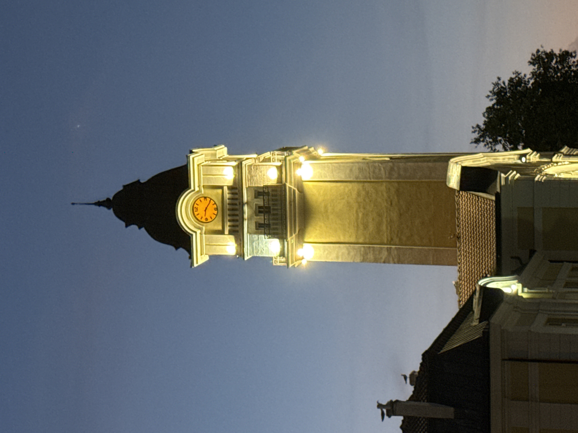 The image shows a clock tower at dusk in Burgas, Bulgaria. The tower is illuminated by artificial lights, highlighting its architectural details, including a clock face showing approximately 12:10. The sky is a clear, deep blue, transitioning to a lighter shade near the horizon. In the foreground, parts of other buildings with tiled roofs are visible, one of which has a prominent chimney with two birds perched on it. A dark tree is visible to the right of the clock tower, its branches silhouetted against the sky. The scene depicts an urban setting at twilight, with the clock tower serving as a focal point.