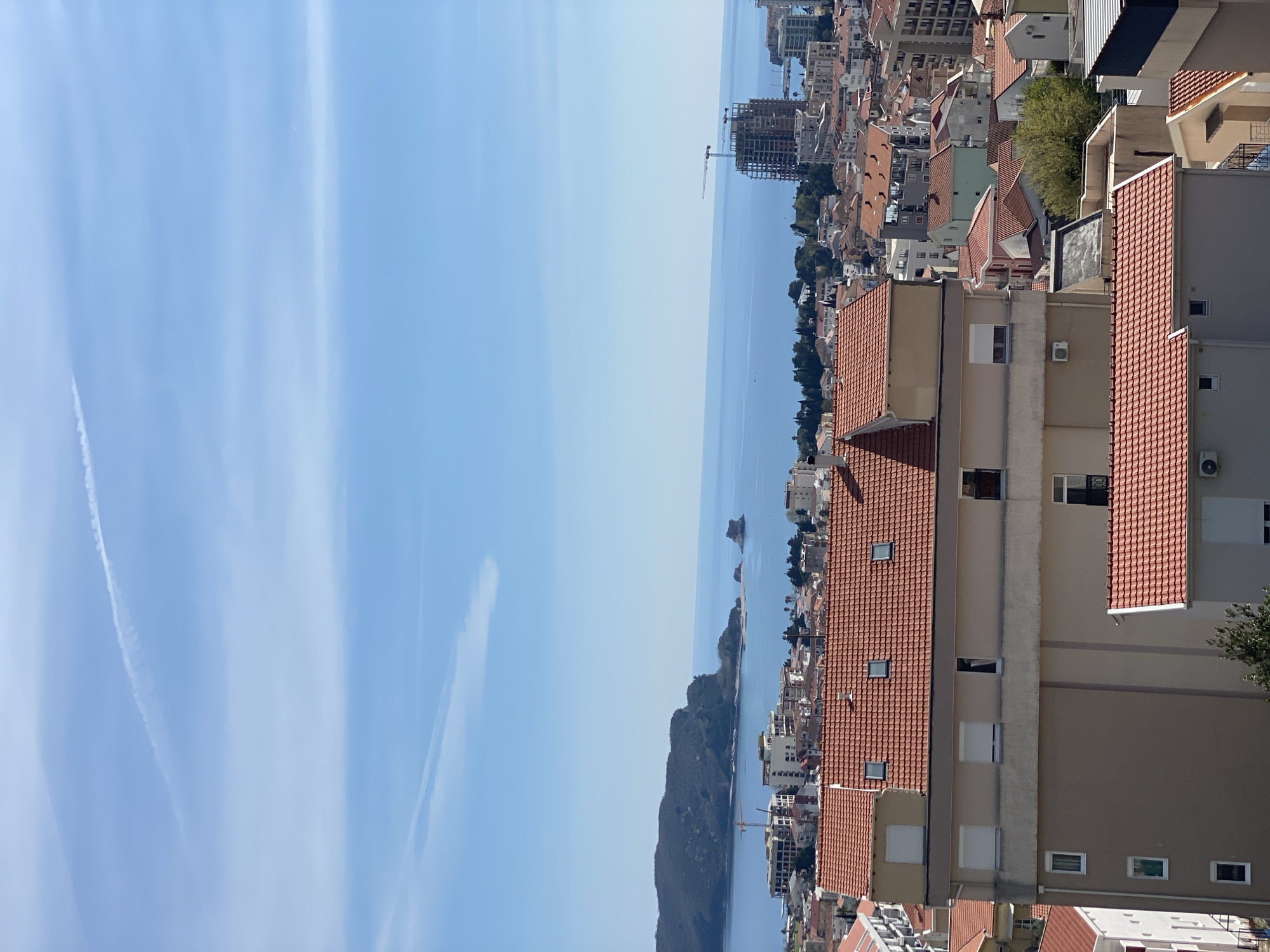The image presents a daytime cityscape, likely Budva, Montenegro, featuring a coastal view and various buildings with prominent red-tiled roofs. The sky is a clear, light blue with faint contrails. The Adriatic Sea extends from the bottom to the top of the frame. Along the right side of the image, buildings are stacked vertically, a mix of residential and commercial structures. In the distance, there is a modern building under construction, evident by a crane. An island or peninsula protrudes into the sea.