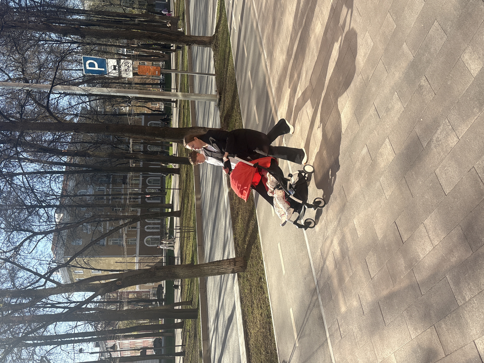 The image shows a woman and a child pushing a stroller on a pedestrian walkway next to a road. The woman is wearing a dark suit, and the child appears to be holding onto the stroller. The stroller has a red cover. The walkway is paved with rectangular stones. The road has white lines painted on it. Trees line the road and walkway. A parking sign is visible in the background. The image was taken during the day with sunlight casting shadows. Based on visual similarities, the location is likely in Perm, Russia.