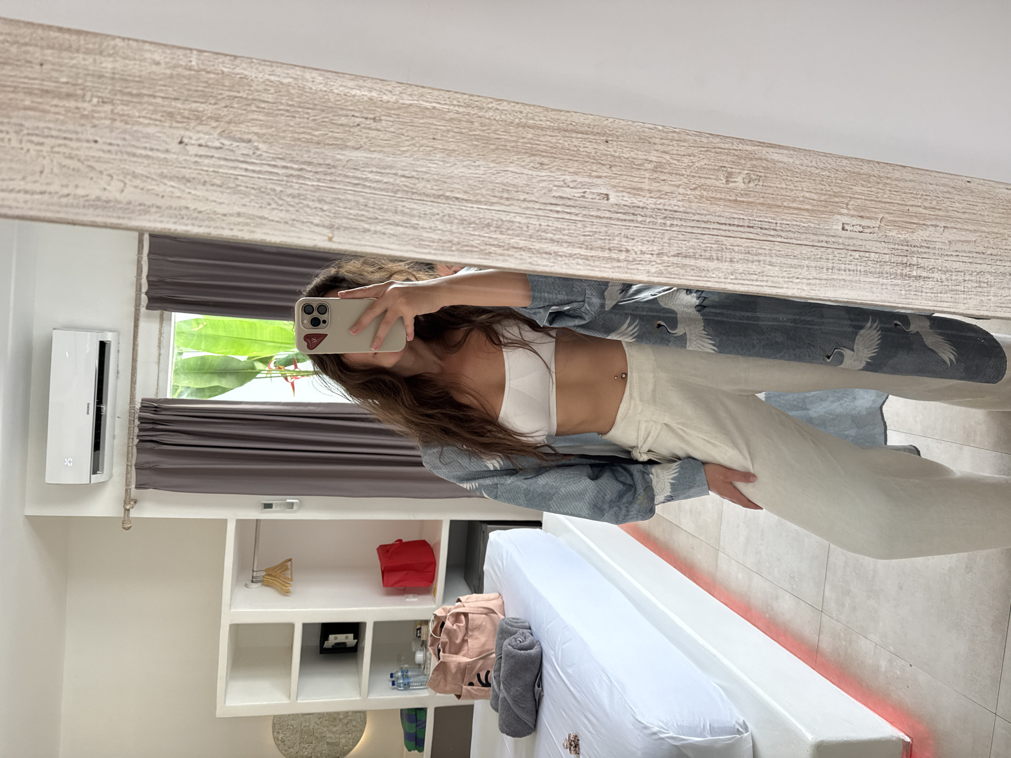 A mirror reflection shows a woman in Unknown, Indonesia, wearing a white crop top and light-colored pants with a blue patterned cardigan, holding a cell phone in front of her face. The room contains a window with brown curtains, an air conditioner, shelving with items, and a bed.