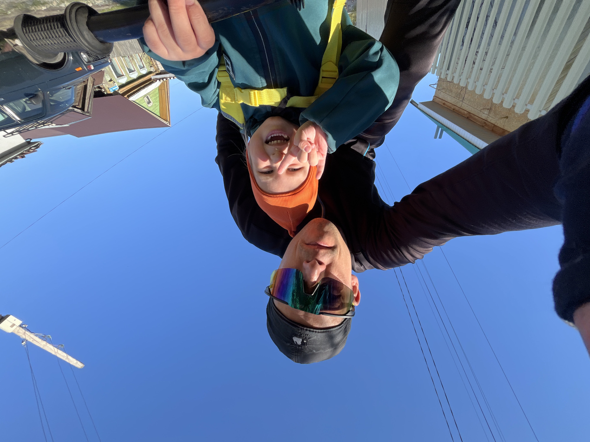 The image, taken from a low angle, shows a man and a child against a clear blue sky. The man is wearing a black cap and reflective sunglasses. The child, wearing a teal jacket with yellow straps and an orange knit hat, is positioned above the man, likely being held. The background includes rooftops and utility poles with wires. Based on visual cues, the photo may have been taken in Chaikovsky, Russia.