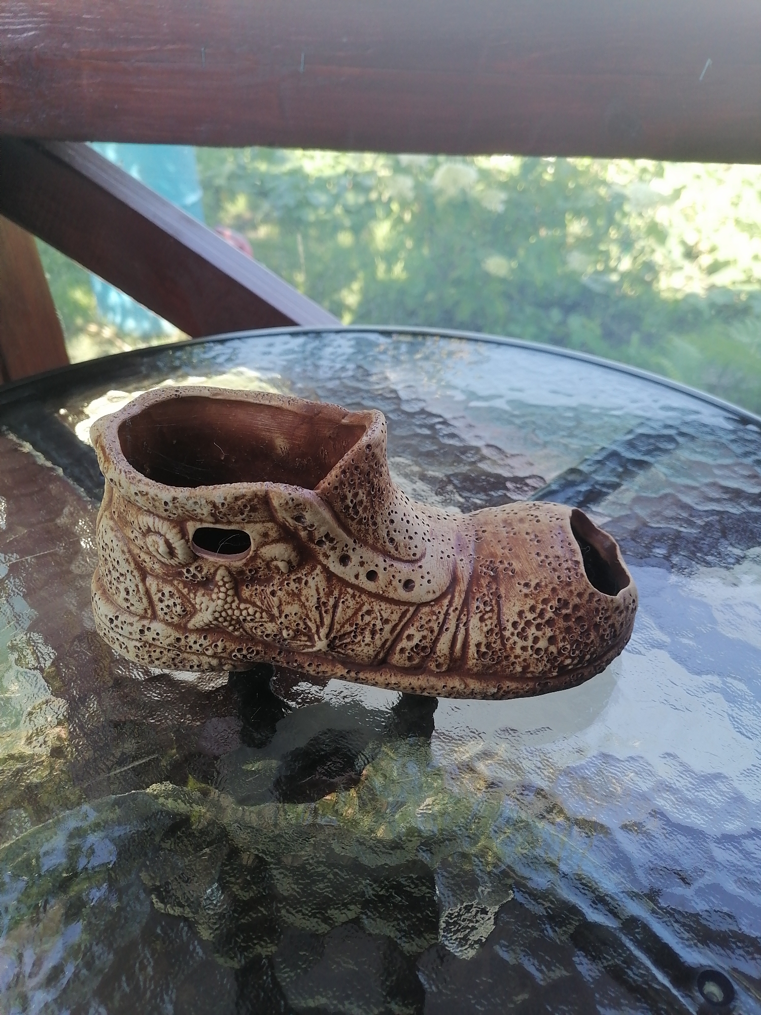 The image depicts a ceramic planter shaped like a shoe, placed on a glass-topped table outdoors. The planter is detailed with perforations and textured patterns resembling the stitching and design of a shoe. The table is situated on a wooden deck or balcony, with lush green foliage and trees visible in the background, suggesting a natural, possibly residential setting. The lighting indicates it is daytime, with sunlight filtering through the leaves and reflecting off the glass table surface. There are no people visible in the image. The location is identified as Pecherniki, Russia, a detail not visually verifiable from the provided image itself.