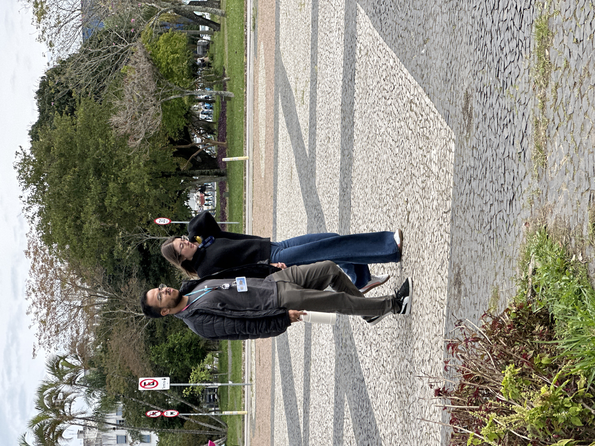 Two individuals, a man and a woman, are walking on a cobblestone path outdoors, with a park-like setting in the background. The man, on the left, wears glasses, a black puffer jacket over a grey t-shirt, and dark pants. He carries a white cylindrical object in his right hand. The woman, on the right, has glasses and wears a black long-sleeved top and wide-leg blue jeans. They are walking side-by-side, appearing to be engaged in conversation or simply walking together.
The background shows palm trees, other deciduous trees with sparse foliage, and buildings. Several traffic signs are visible on the left, including a red circle with a horizontal bar and a downward-pointing arrow, and another with a red circle and a downward-pointing arrow with a vehicle symbol. A circular sign with "30" inside a red circle is visible further back. The ground is a distinctive patterned cobblestone walkway, with some areas of grass and plants visible in the foreground and to the right. The overall scene suggests a public outdoor space in an urban environment, likely during the day under an overcast sky. The location is the Beira-Mar Norte avenue in Florianópolis, Brazil, a prominent waterfront promenade known for its public spaces and recreational areas.