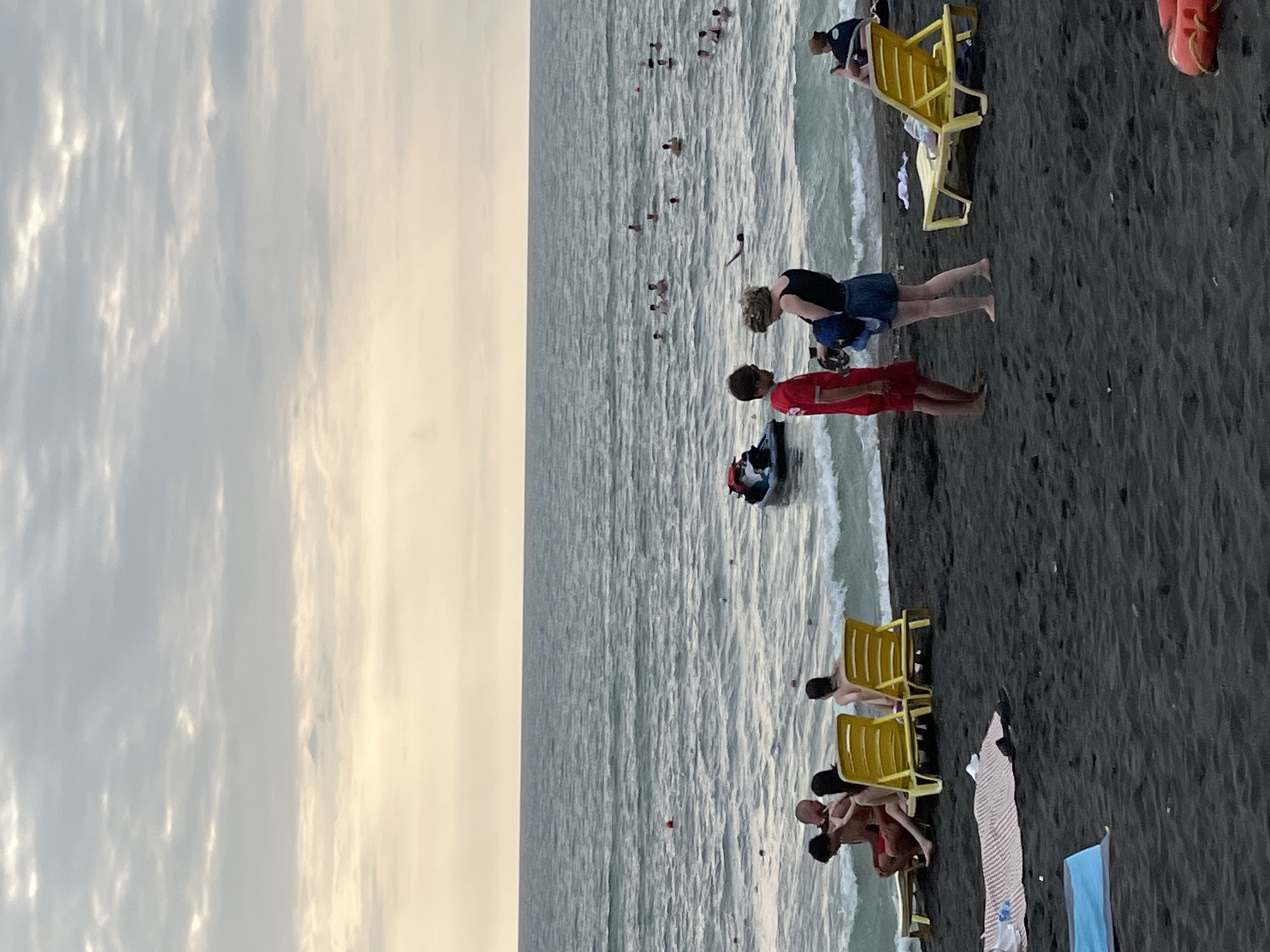 The image depicts a beach scene in Ureki, Georgia, characterized by its distinctive black sand. In the foreground, several individuals are present on the dark sand. A group of people are seated on bright yellow lounge chairs near the water's edge, with some of them facing the sea. Further inland, a patterned blanket is laid out on the sand, with a blue mat nearby. Two individuals, a male in a red shirt and shorts and a female in a black tank top and denim shorts, are standing and interacting on the beach. A yellow lounge chair is positioned behind them. In the middle ground, the sea is visible, with numerous people swimming and enjoying the water. A jet ski is also present in the water. The background shows a tranquil sea under a cloudy sky with a hint of a sunset glow.