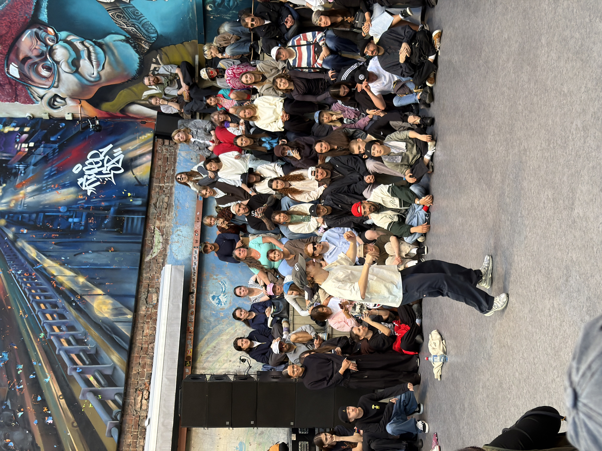 The image depicts an outdoor event with a large group of people gathered in what appears to be a public space, possibly a courtyard or an open-air venue in Saint Petersburg, Russia. The background features a large, vibrant mural on a brick wall, showcasing a stylized depiction of a bridge and a cartoonish caricature of a person's face with oversized glasses.
In the foreground, a young person is actively engaged in a dance, their body in mid-motion, wearing dark pants and white sneakers. They are positioned on a gray, textured floor. Surrounding the dancer is an audience seated on wooden tiered risers. The audience members are diverse in age and gender, dressed in casual, contemporary clothing. They are observing the performance, some with their phones out.
To the left of the scene, several large black speaker cabinets are stacked, indicating a possible musical performance or event. The overall atmosphere suggests a cultural gathering, potentially a street performance, festival, or a dance battle. The lighting suggests it is daytime. The art style of the mural and the casual attire of the attendees point towards a modern, urban setting.