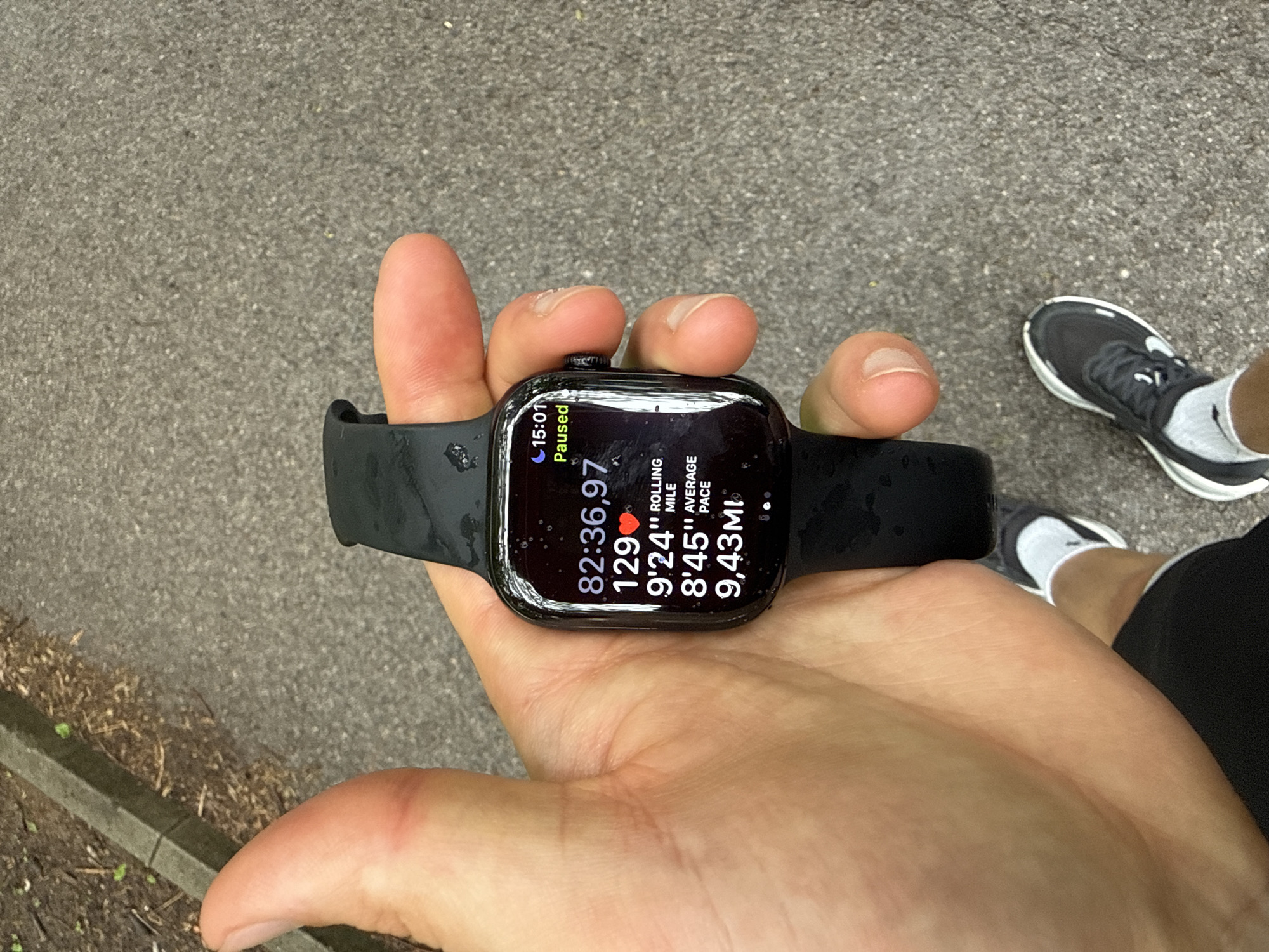 A person is holding a black Apple Watch in their hand, displaying the paused workout metrics. The watch face shows a time of 15:01, with the workout paused. The duration of the workout is 82 minutes, 36.97 seconds. The heart rate is 129 beats per minute. The rolling mile is 9 minutes, 24 seconds, and the average pace is 8 minutes, 45 seconds. The distance covered is 9.43 miles. The watch and the hand holding it appear to be damp, possibly from sweat or rain. The hand is Caucasian. A portion of the person's leg is visible, showing a running shoe, a white sock, and black shorts. The background is an asphalt surface and some greenery. This image was not taken in Sofia, Bulgaria.