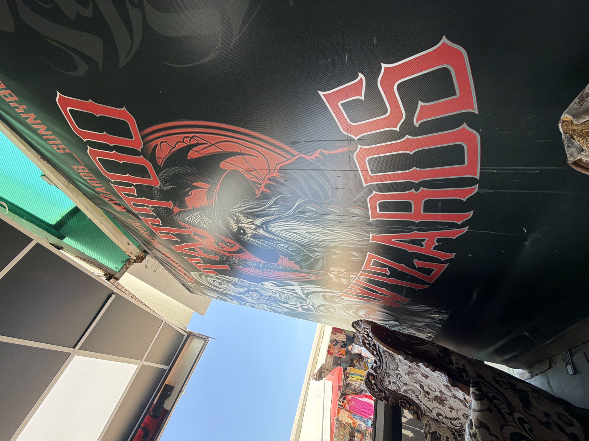 The image shows a close-up of a black banner with red lettering and a drawing of a wizard. The text on the banner reads "TATTOO WIZARDS SUNNY B" with "WIZARDS" more prominently displayed at the bottom. The drawing depicts a wizard with a long white beard, a pointed hat, and a stern expression, surrounded by a red circular design and what appear to be dragon wings or a cloak. The banner is tilted and appears to be attached to a structure with a green corrugated plastic roof. In the background, out of focus, there are stalls with colorful clothing and a bright blue sky. The scene suggests an outdoor market or a stall selling tattoo services. Based on the visual cues, it is likely located in the historic town of Nesebar, Bulgaria, possibly within the Old Town area known for its vibrant market stalls. A portion of an ornate, upholstered chair is visible in the lower left foreground.