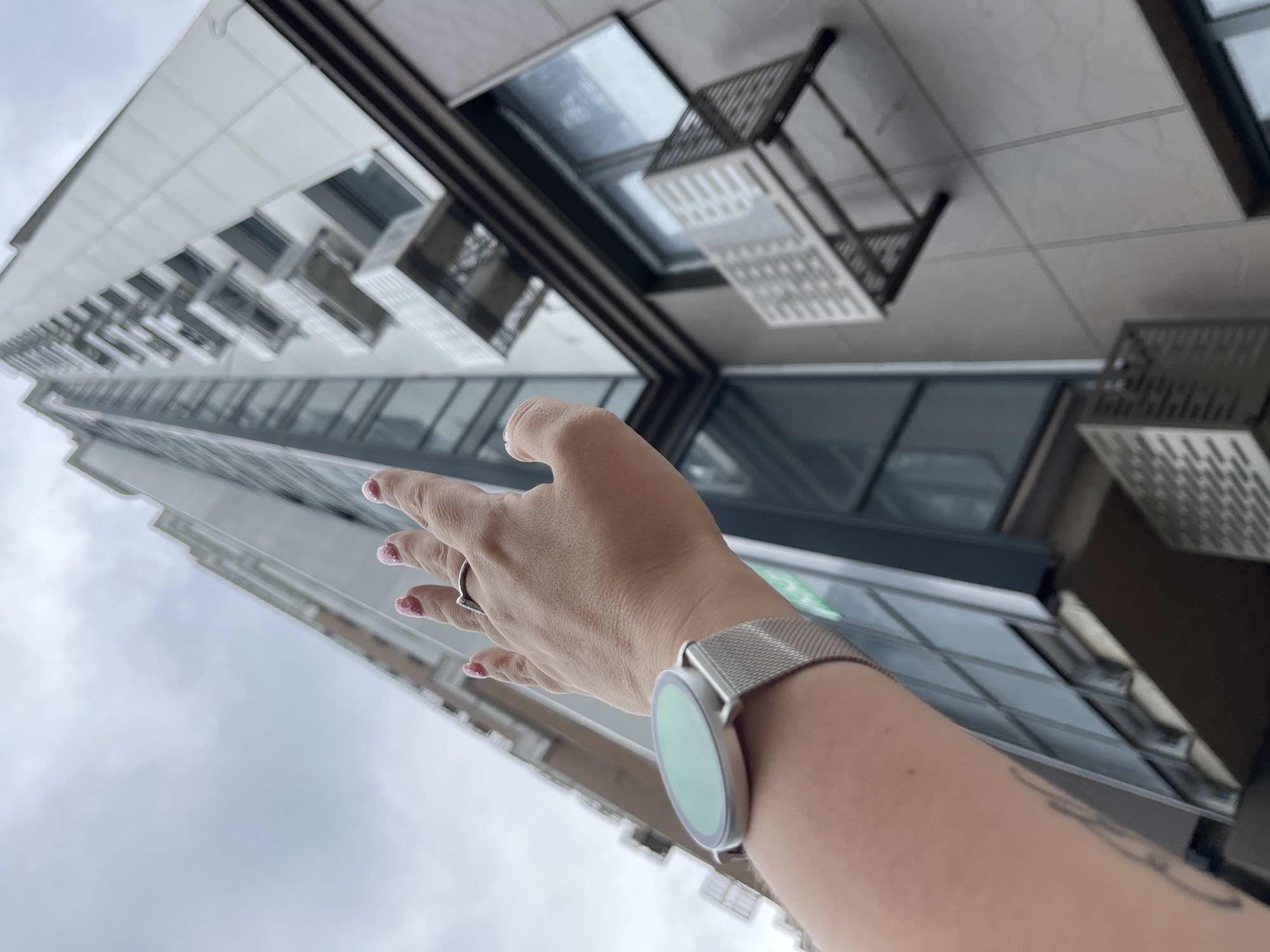 A low-angle, outdoor shot focuses on a person's outstretched hand with a smartwatch on their wrist, reaching towards a tall, modern apartment building. The building, constructed with white and grey panels, features numerous balconies and large glass windows. Some windows have air conditioning units mounted outside. The sky above is overcast. The scene is likely in Sochi, Russia, a city known for its modern architecture and coastal location.