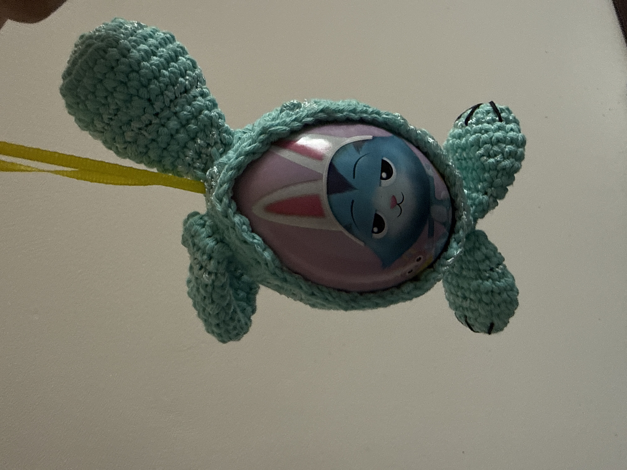 The image shows a crocheted teal bunny ornament hanging by a yellow ribbon against a plain, light-colored background. The bunny has large, floppy ears and a hooded head that frames a colorful oval object. This oval object is adorned with a cartoonish blue cat character wearing bunny ears on a pink background. The cat has a mischievous expression with narrowed eyes and a small pink nose. The crocheted bunny also has two dangling "feet" at the bottom, each with two black stitches resembling toes. The lighting suggests an indoor environment. The image does not provide any information about the city of Stara Zagora, Bulgaria.