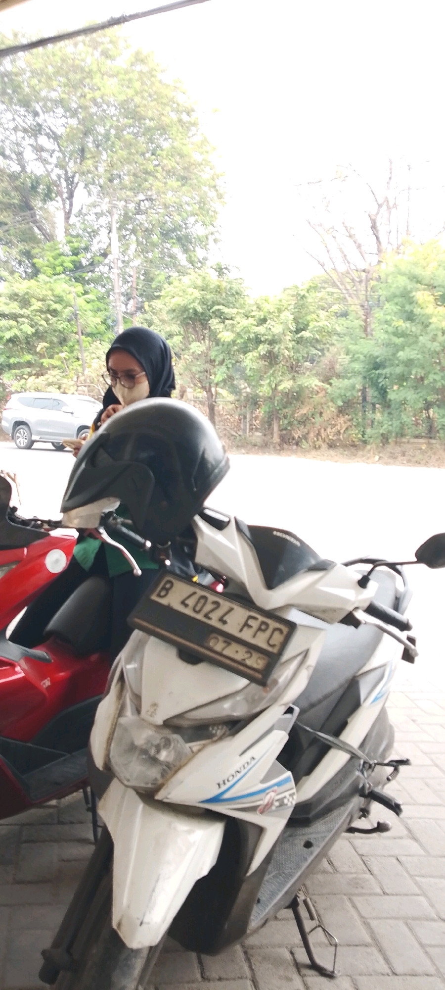 A person wearing a black hijab and a light-colored face mask stands near a red motorcycle in front of a white scooter. The person appears to be looking down at their hands, which are holding a smartphone. A black helmet rests on the handlebars of the scooter. The license plate on the scooter reads "B 4024 FPC". The background shows a line of trees and a silver SUV parked on a paved road. The scene is set in an outdoor environment, likely a roadside or parking area, with a tiled floor. The overall lighting suggests it is daytime.