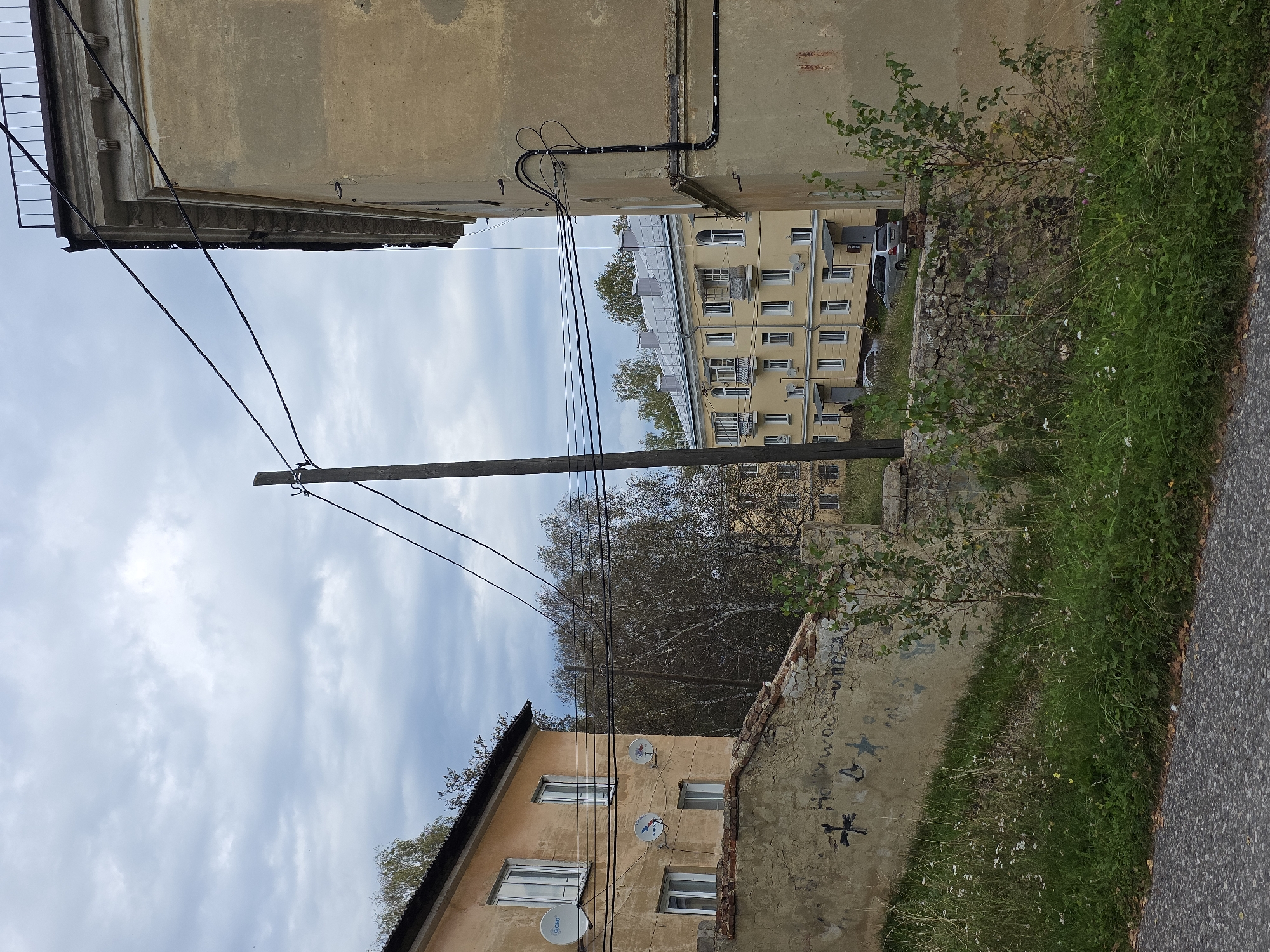 The image depicts an outdoor scene in a city, likely Magnitka, Russia, on a cloudy day. In the foreground, a paved road or sidewalk is visible, bordered by overgrown green grass and weeds with some scattered fallen leaves. A low, weathered stone and brick wall with graffiti runs along the edge of the grassy area. Beyond the wall, a two-story, light yellow apartment building with several windows and satellite dishes on its facade is partially visible. Behind this building, a taller, aged, pale yellow building with decorative architectural details and a metal railing on its roof occupies the right side of the frame. Overhead, a network of black electrical wires stretches across the sky, supported by wooden and metal poles. In the middle ground, another long, pale yellow apartment building with multiple windows and balconies is visible. The sky is overcast with a mix of light and dark gray clouds. Several bare trees with thin branches are interspersed between the buildings, some appearing to be birch trees. A light-colored minivan is parked in front of the middle ground apartment building. No people are clearly visible in the image.